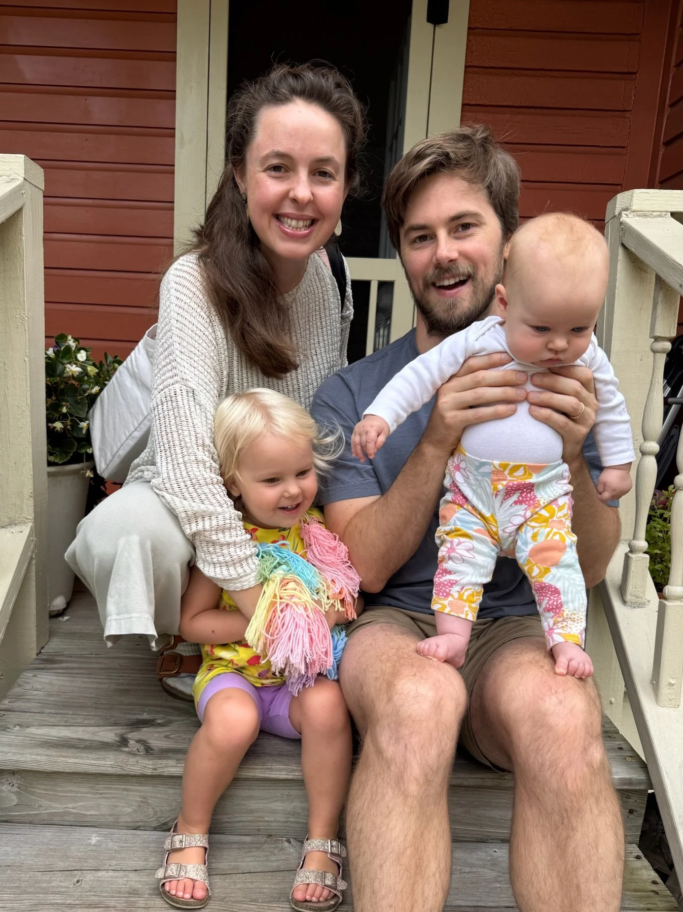 A family of four sitting on a porch steps, smiling. The father is holding a baby, and a young girl is sitting beside him, while the mother leans over, all smiling.