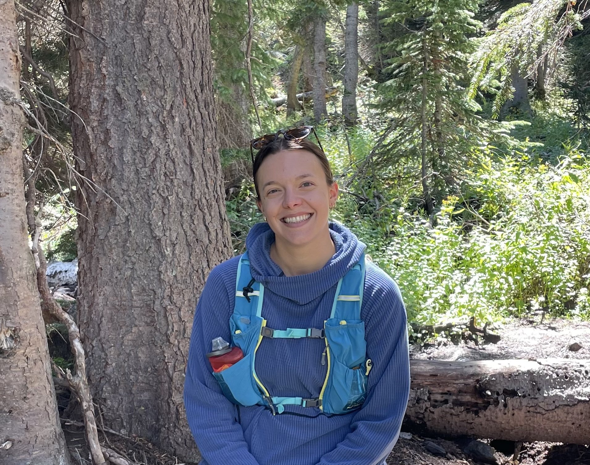 A smiling woman in a blue jacket with a blue hiking backpack outdoors in a forested area next to trees and greenery.