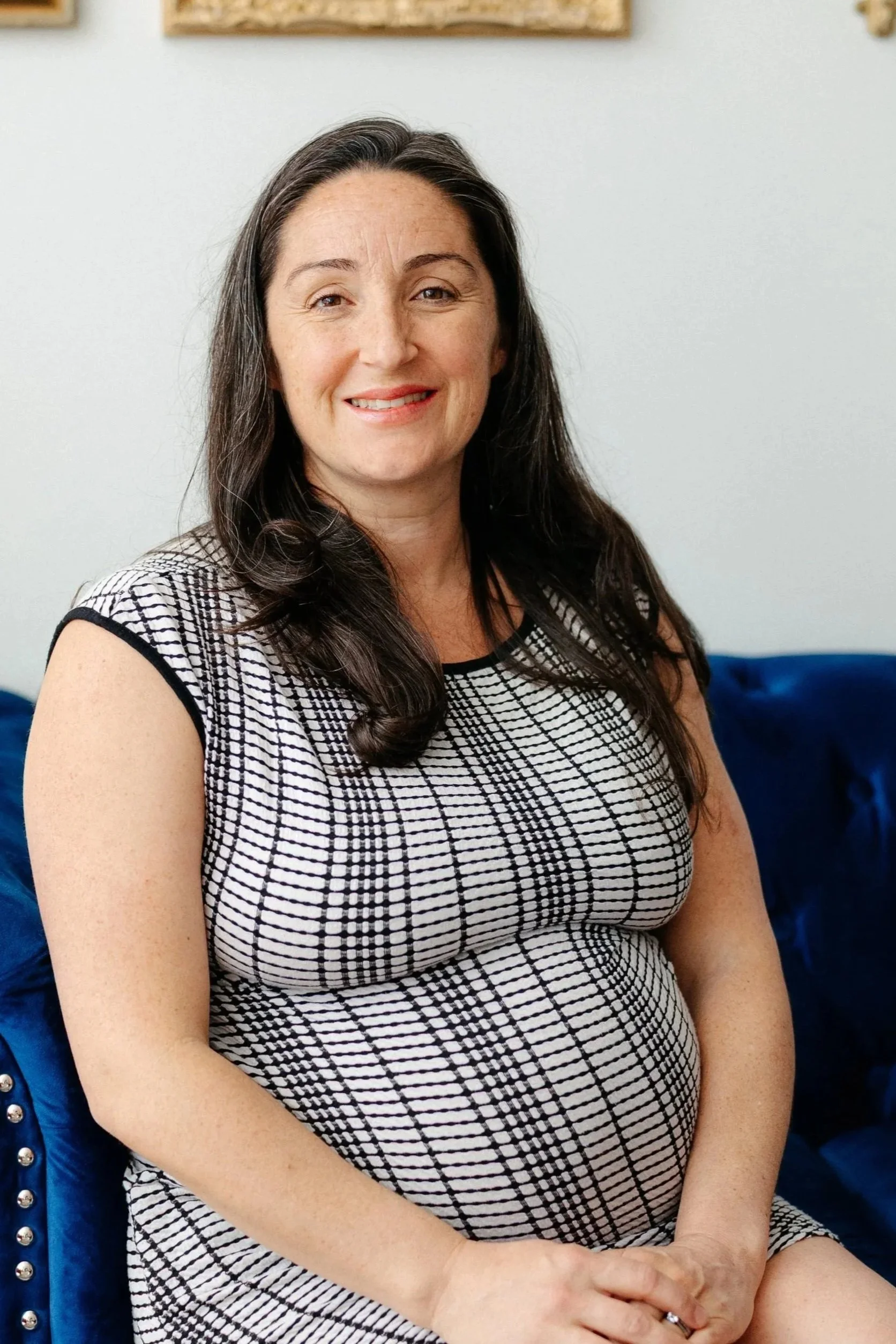 Pregnant woman smiling with long black hair while sitting a blue couch wearing a black and while dress