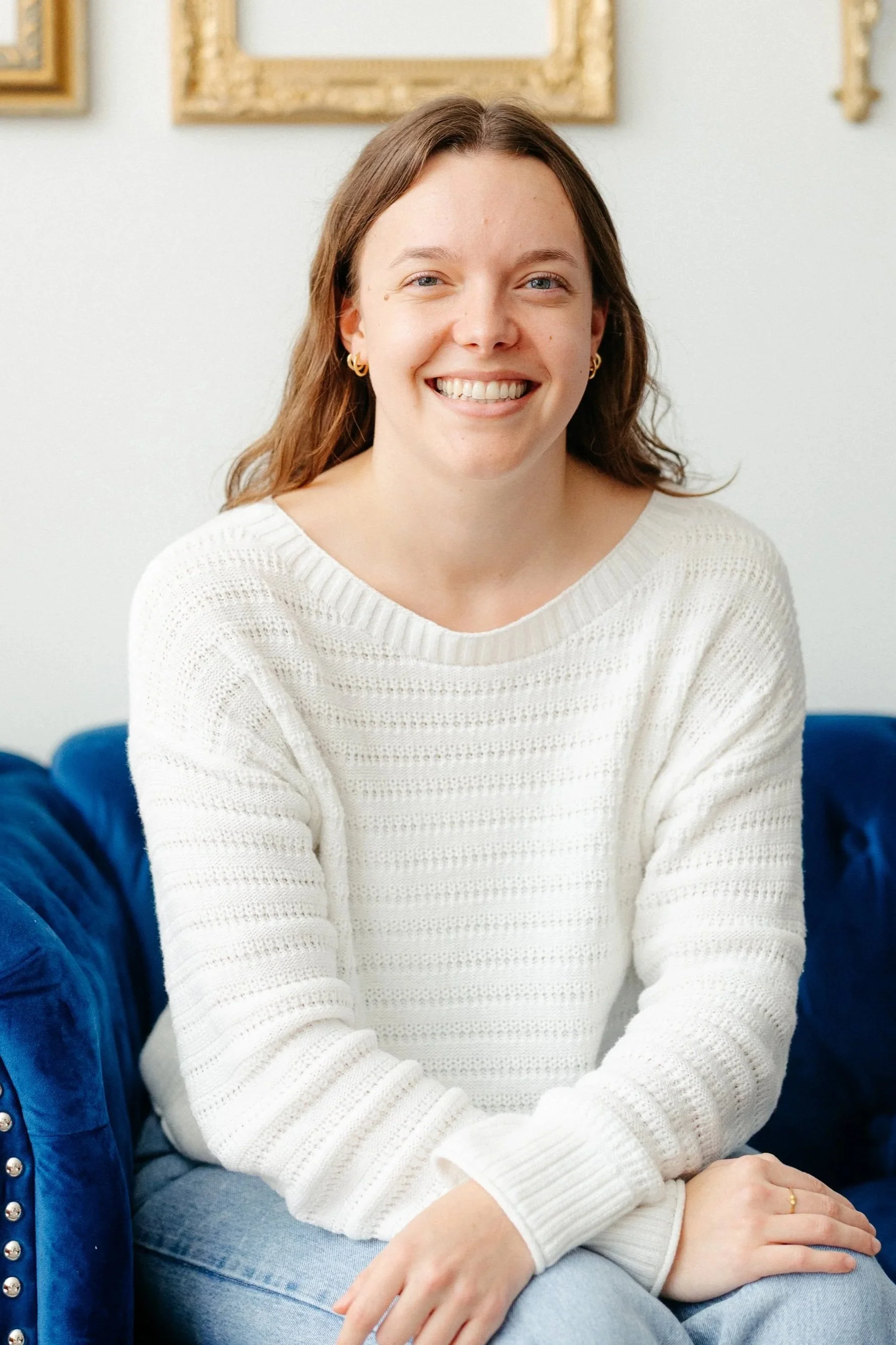 Woman smiling while sitting a blue couch wearing a white sweater and gold earrings
