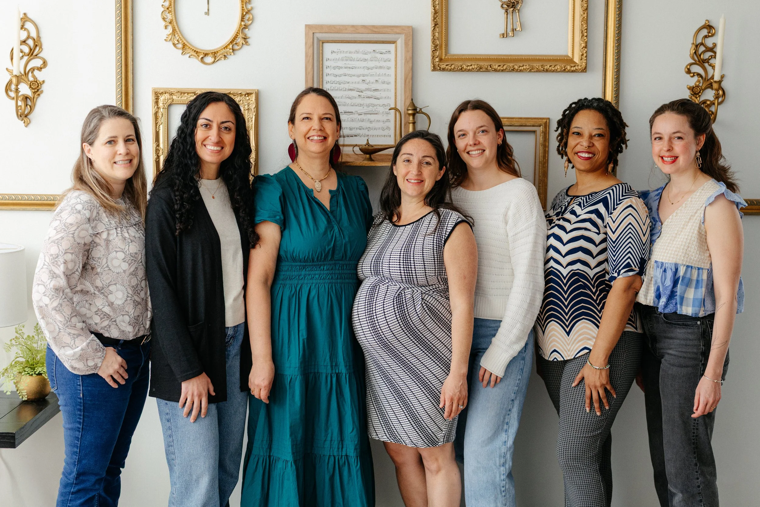 Seven women standing side by side in a room with a white wall decorated with gold picture frames and a sheet of music in a frame. They are smiling and dressed casually.