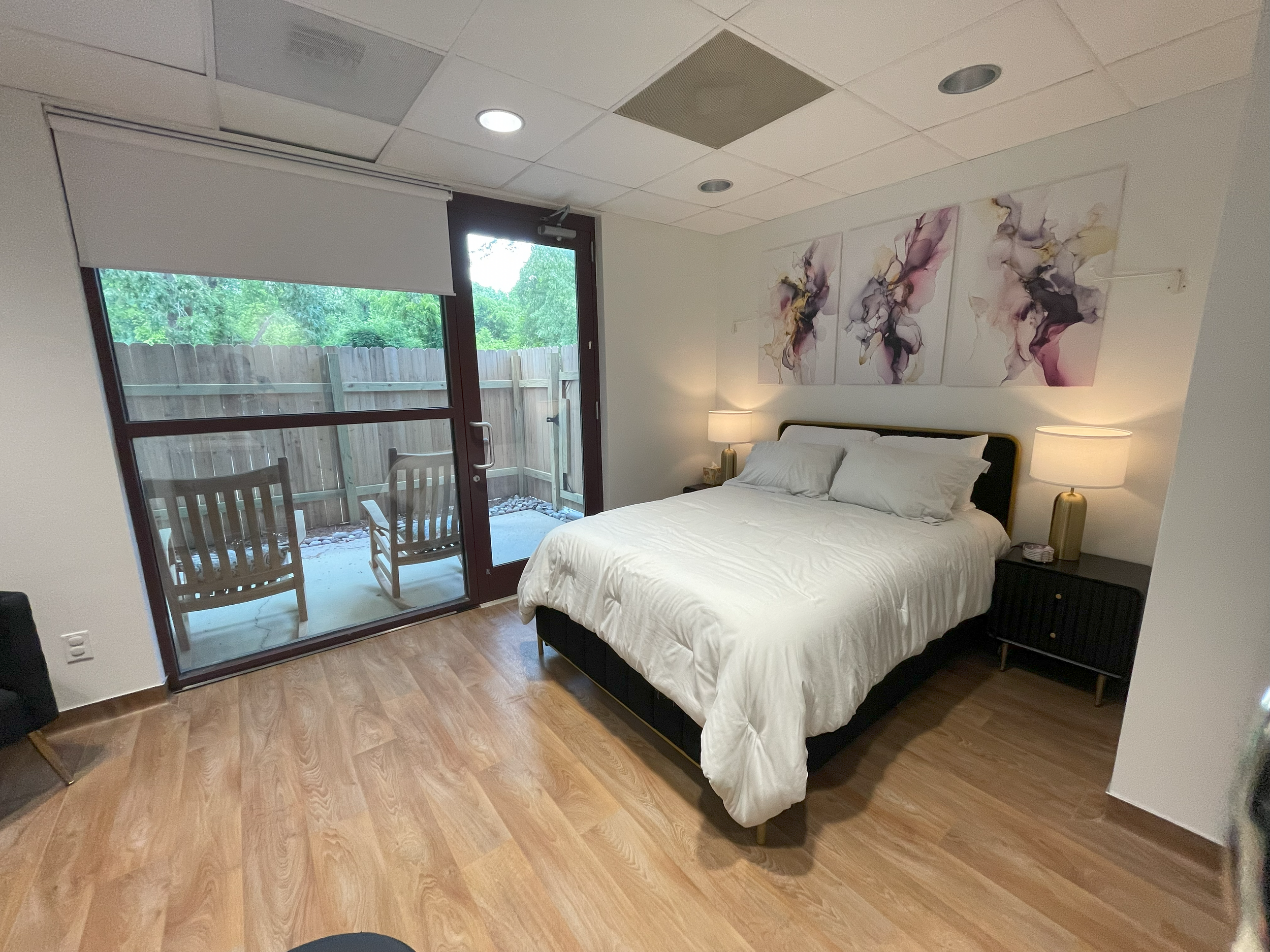 A bedroom with a bed having white bedding and pillows, two bedside lamps, and three abstract flower paintings above the headboard. There is a sliding glass door leading to a small outdoor patio with two chairs and a wooden fence, and a ceiling with r