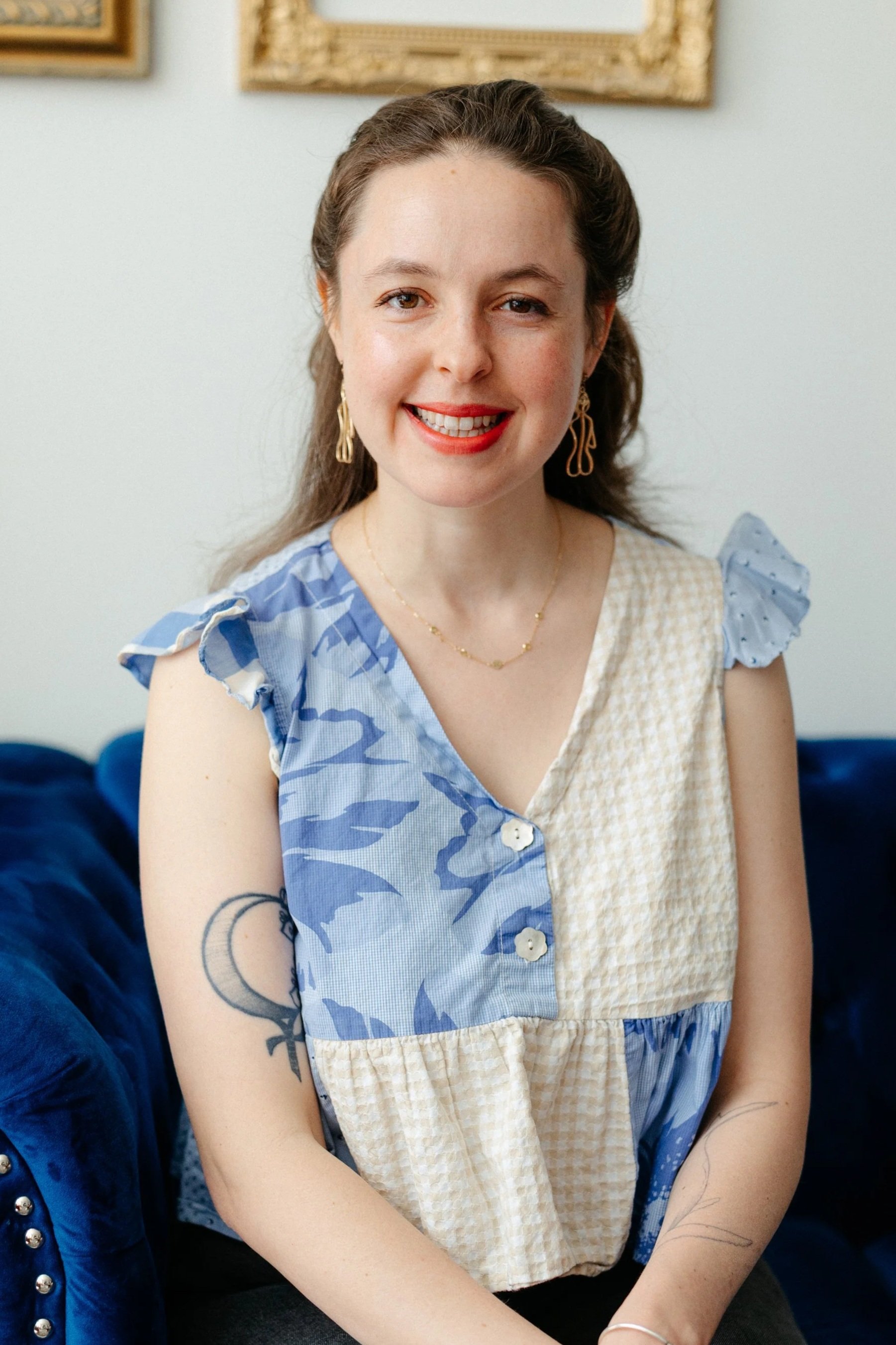 Woman smiling while sitting a blue couch wearing a white and blue shirt and gold earrings