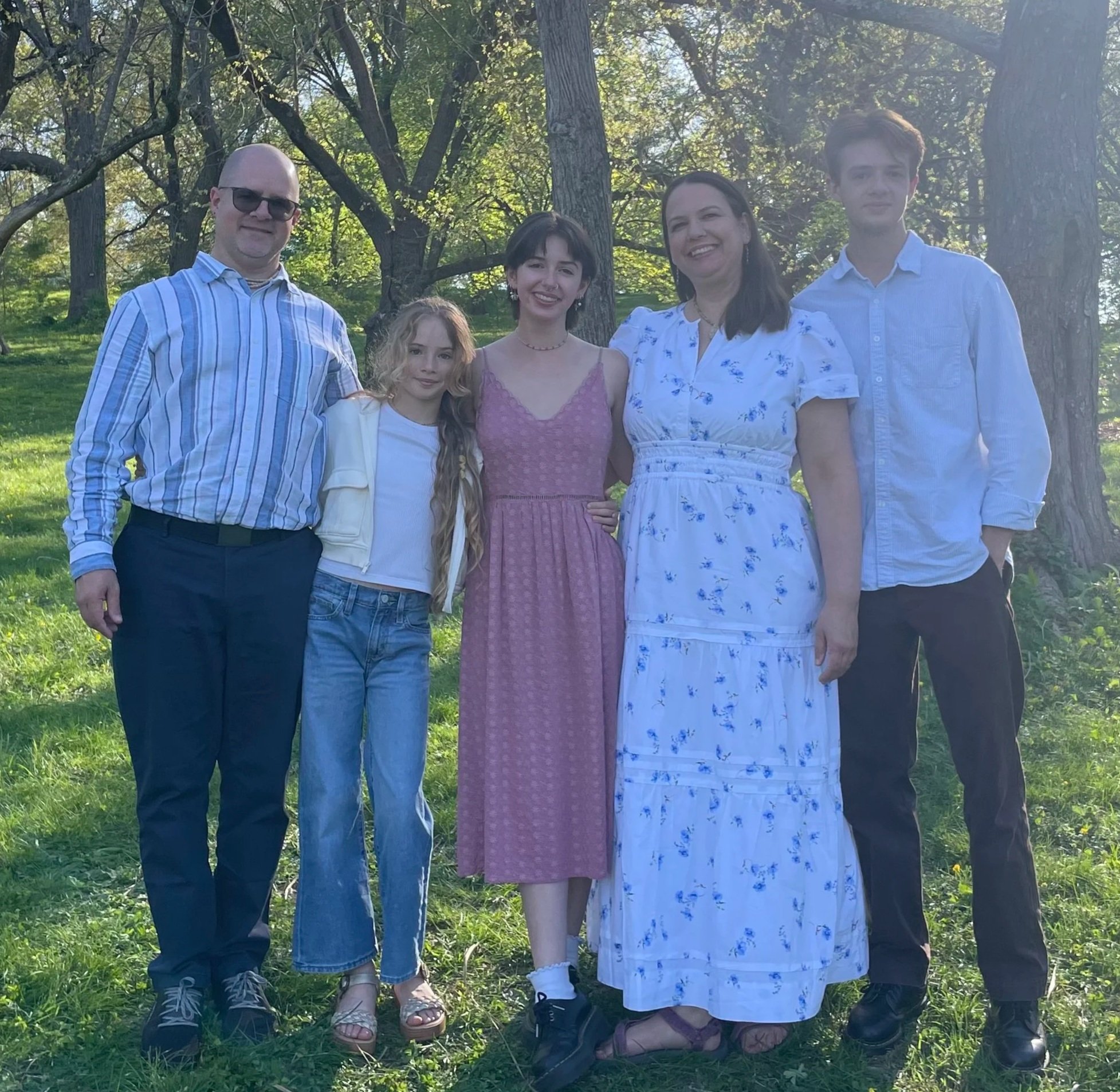 Group of five people standing outdoors on grass in a wooded area, smiling at the camera, with sunlight filtering through trees in the background.