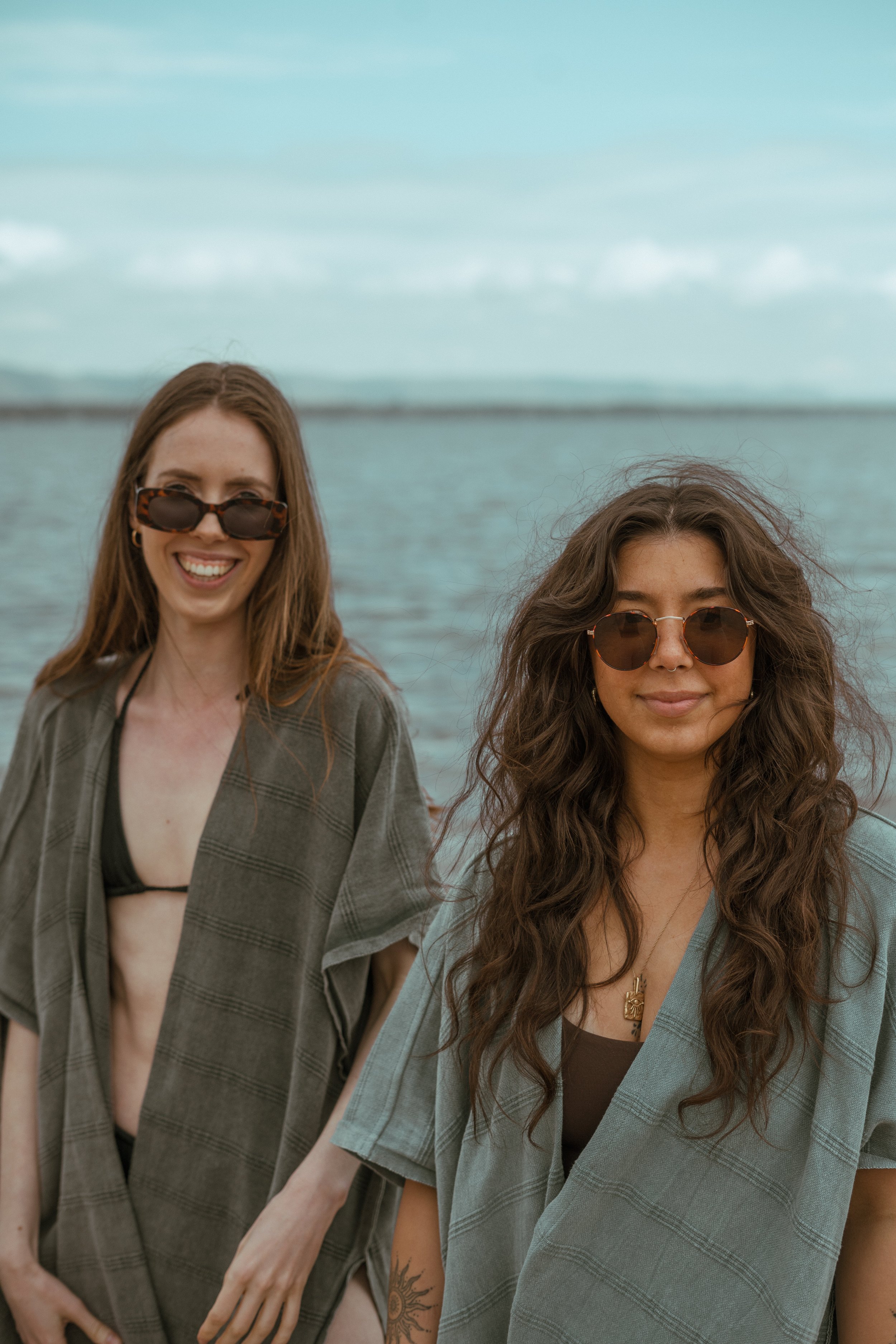Two women wearing sunglasses and beach cover-ups, smiling at the camera with a body of water in the background.