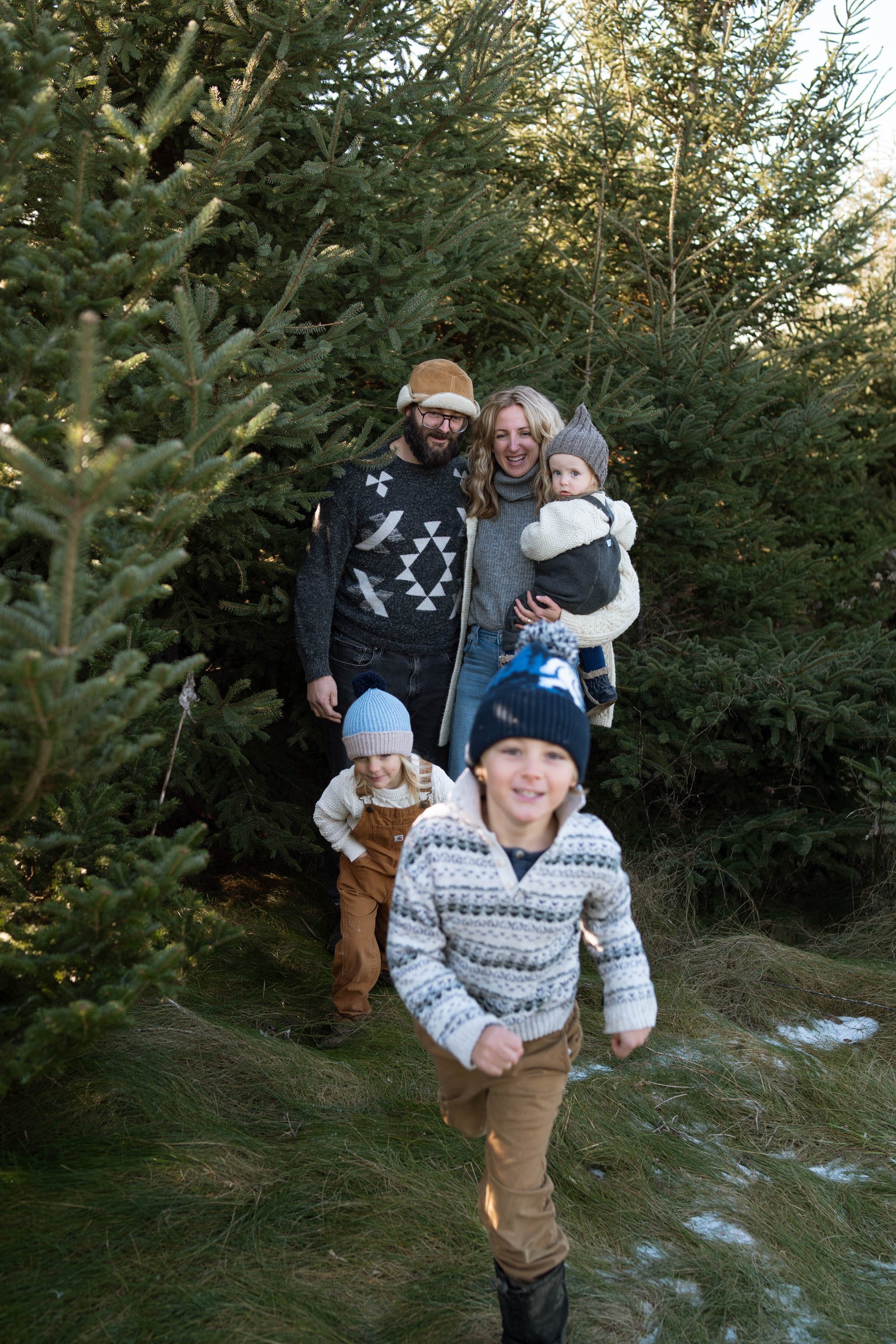 Family in winter clothing in a pine tree forest, with two children playing in the foreground.