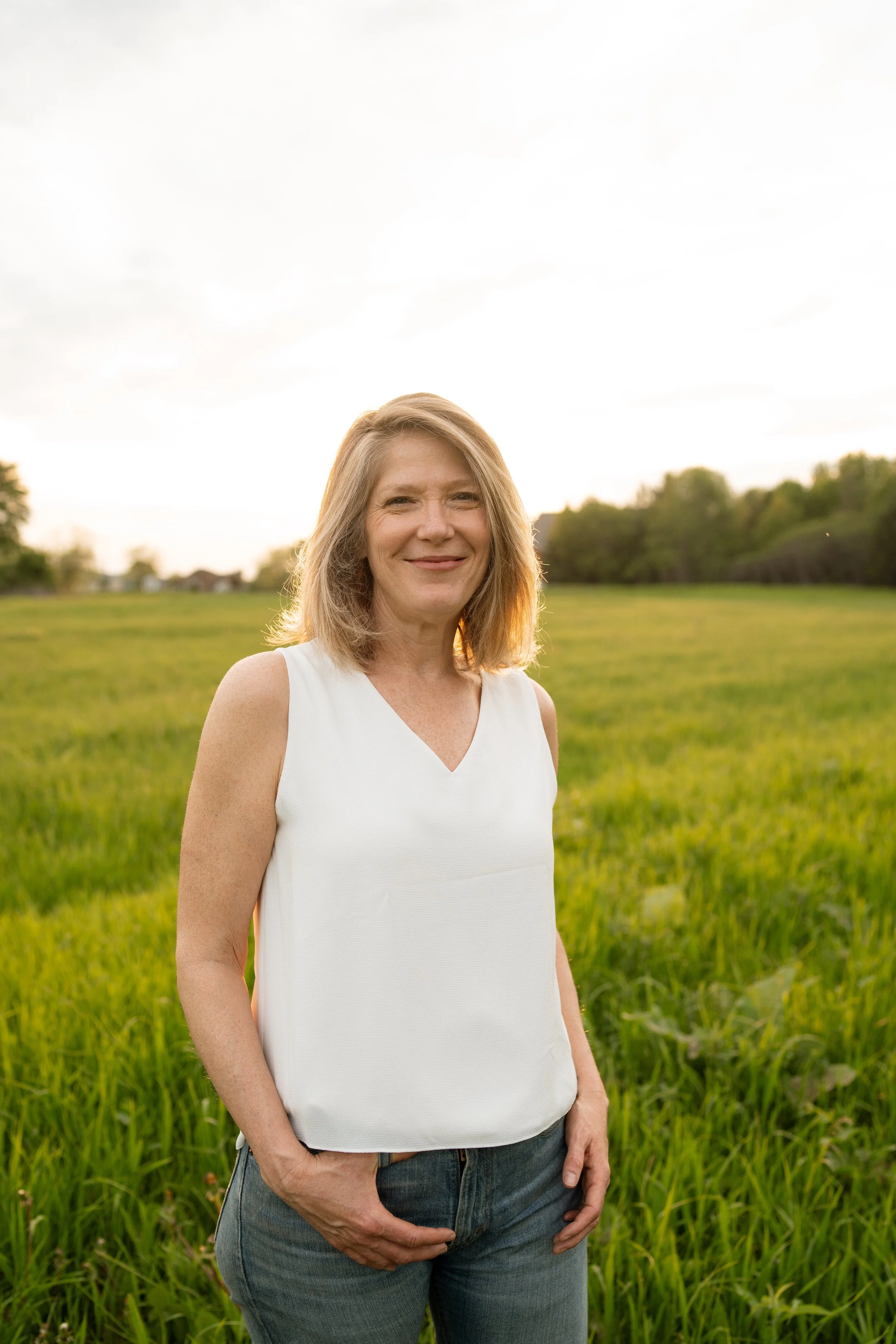 Woman in white tank top stands in a green field under a clear sky.
