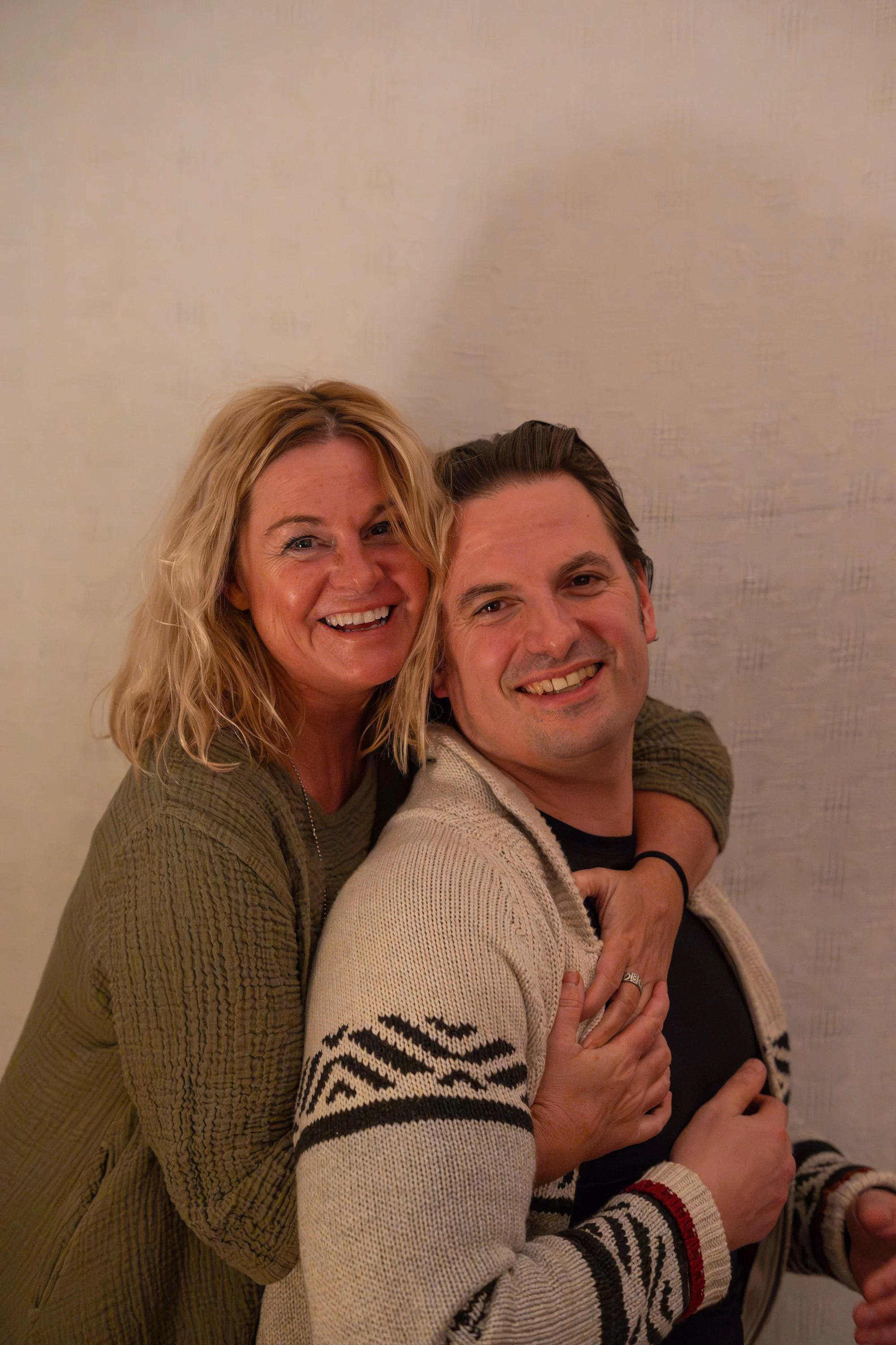 A woman and a man smiling together, embracing, both wearing casual sweaters against a neutral background.