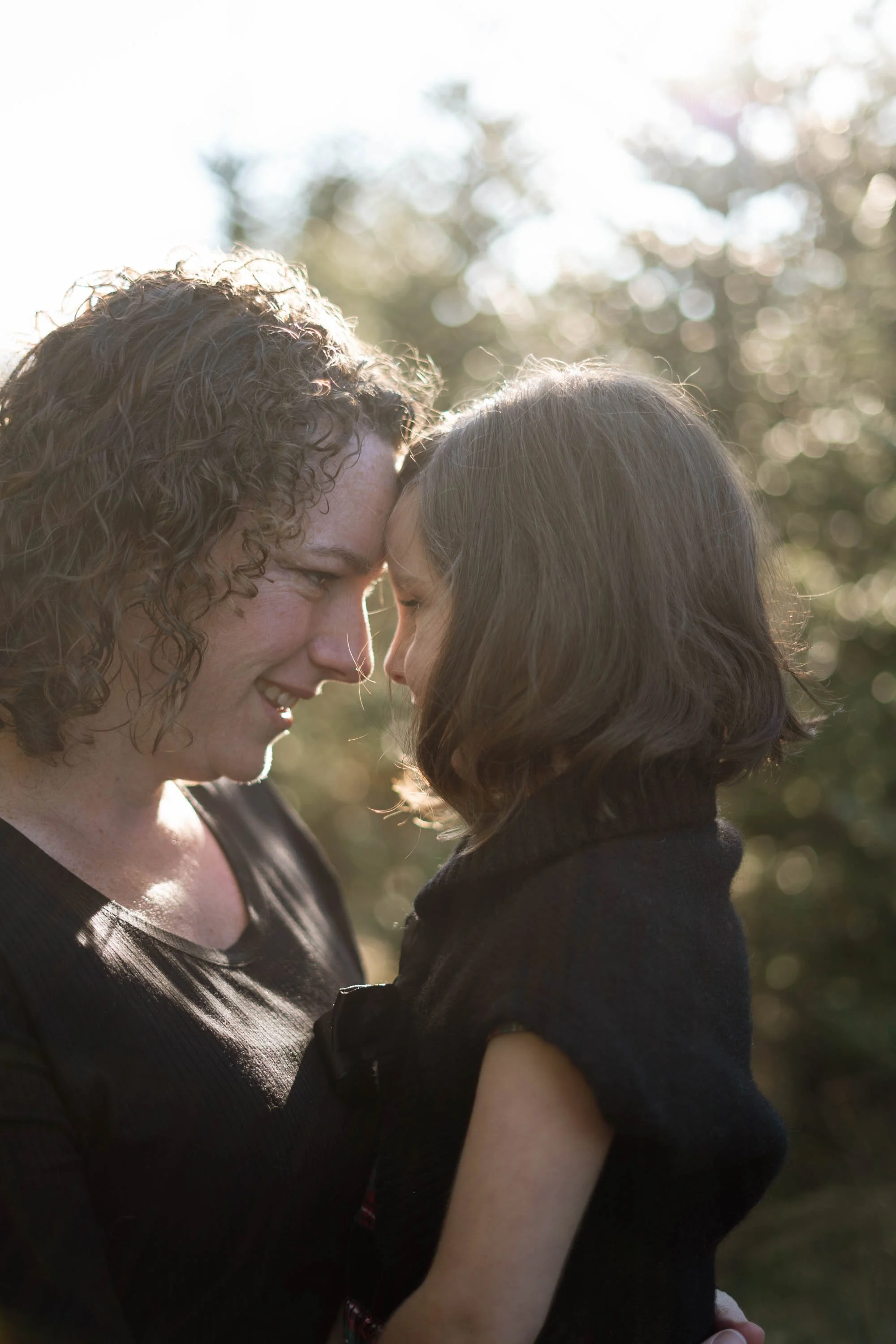 A woman and a child touching foreheads tenderly in a sunlit outdoor setting.