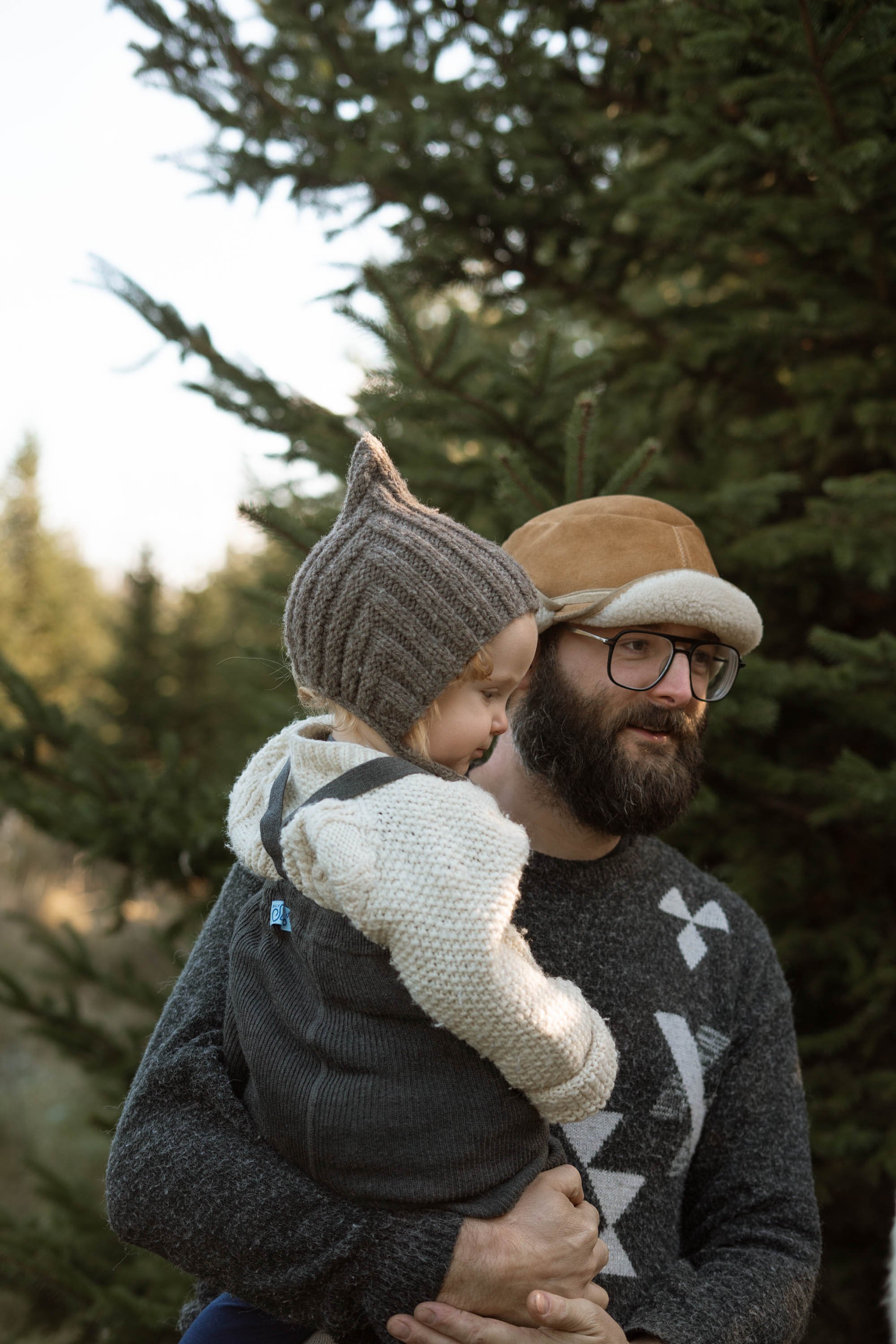 Man holding child outdoors near evergreen trees, both wearing knit hats and warm clothing.
