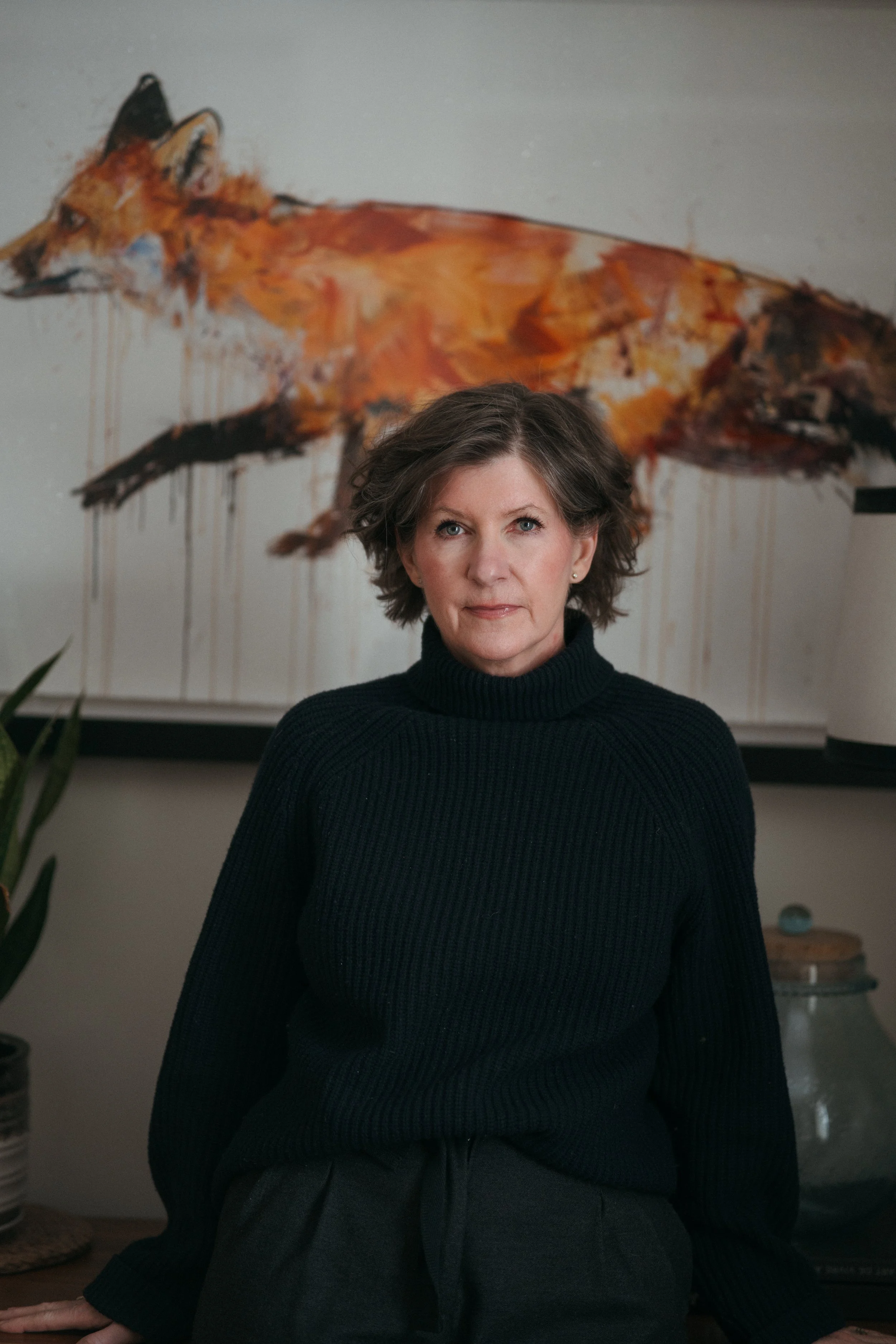 A woman wearing a brown hat and white dress sits barefoot on a white stool on a gravel road surrounded by trees.