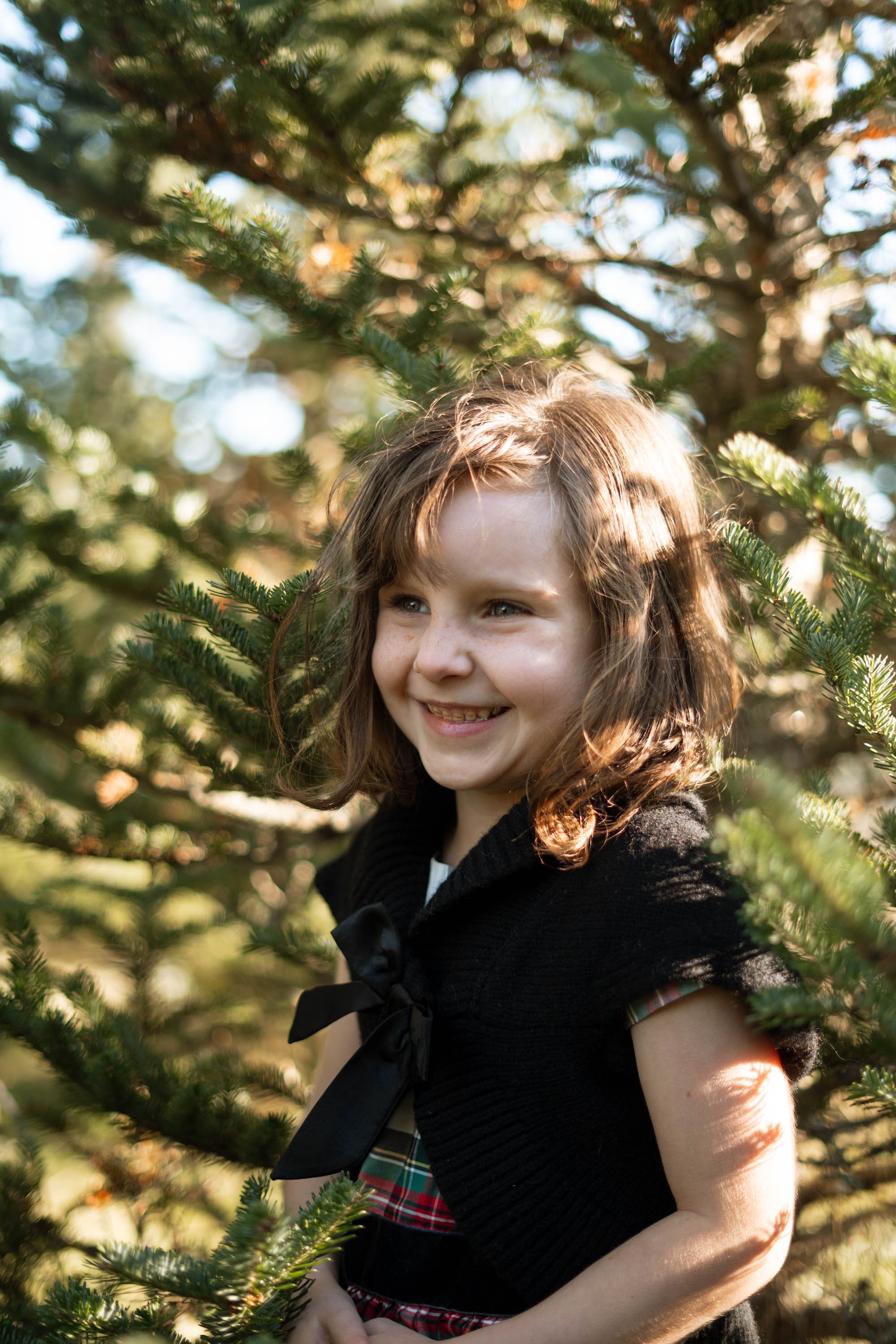 Child smiling among pine tree branches in daylight.