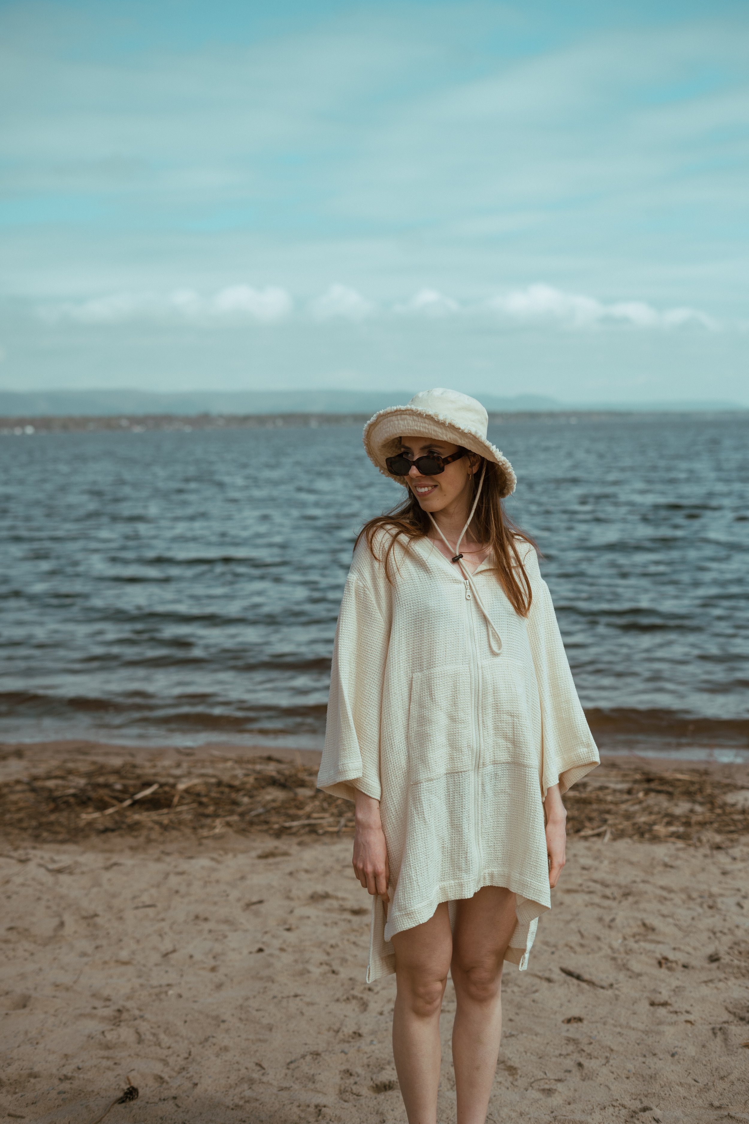 Woman on beach wearing a white cover-up, sunglasses, and a hat, with ocean in background.