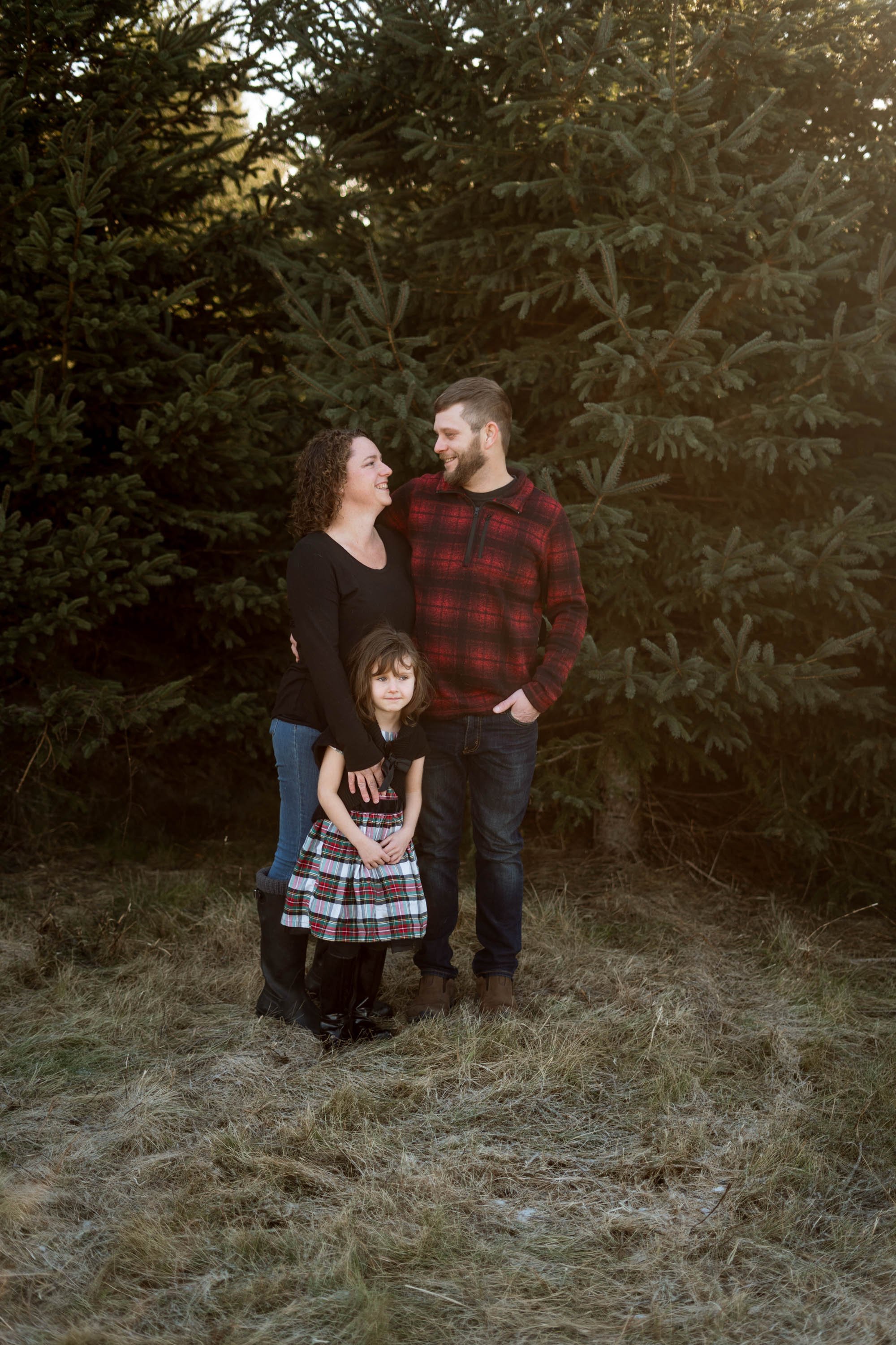 Family posing outdoors in front of evergreen trees, with a woman, man, and child. The woman and man are smiling at each other, and the child stands in the front wearing a plaid skirt.