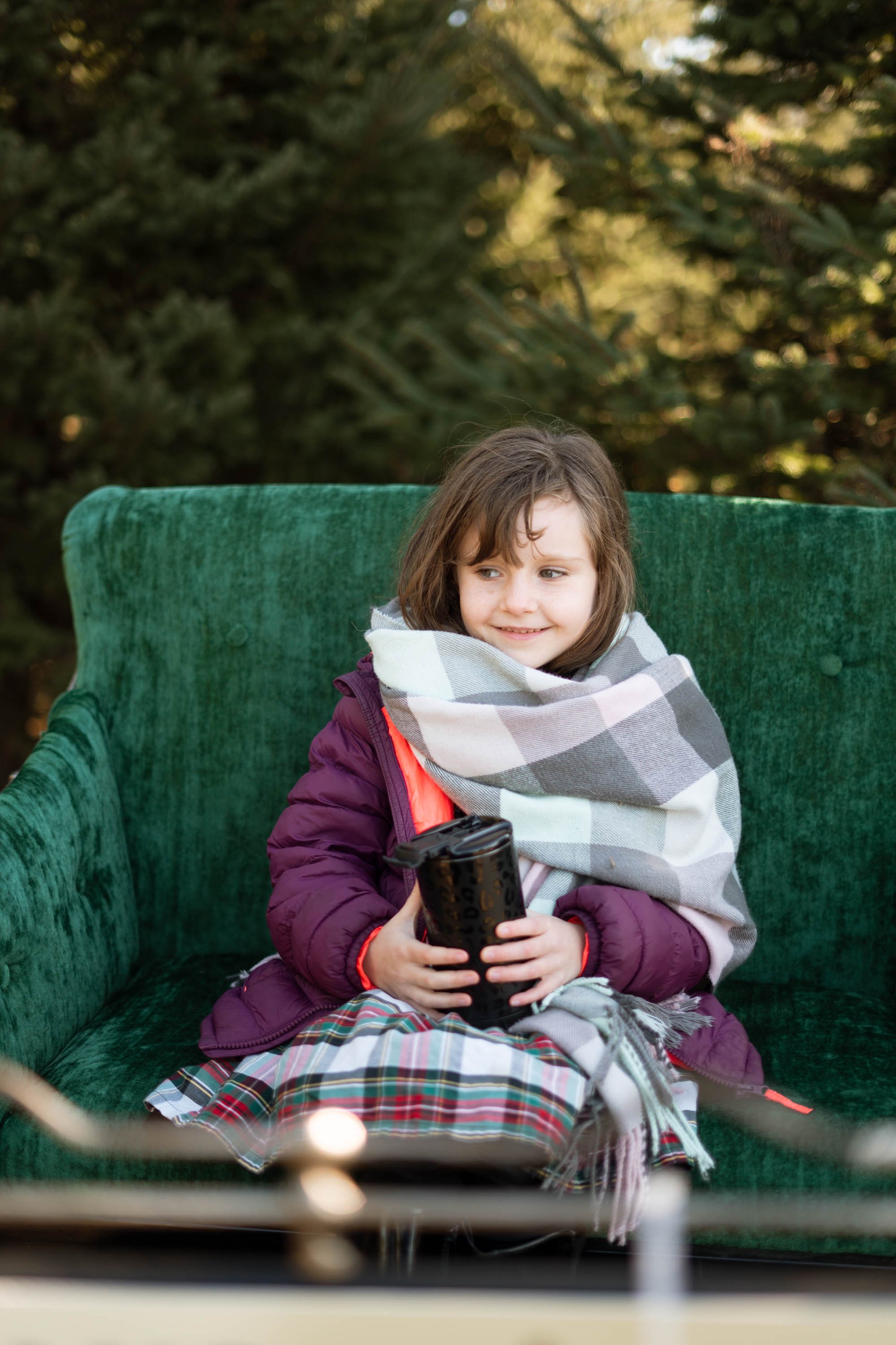Child sitting on a green couch outdoors, wearing a purple jacket and plaid scarf, holding a black mug, with evergreen trees in the background.