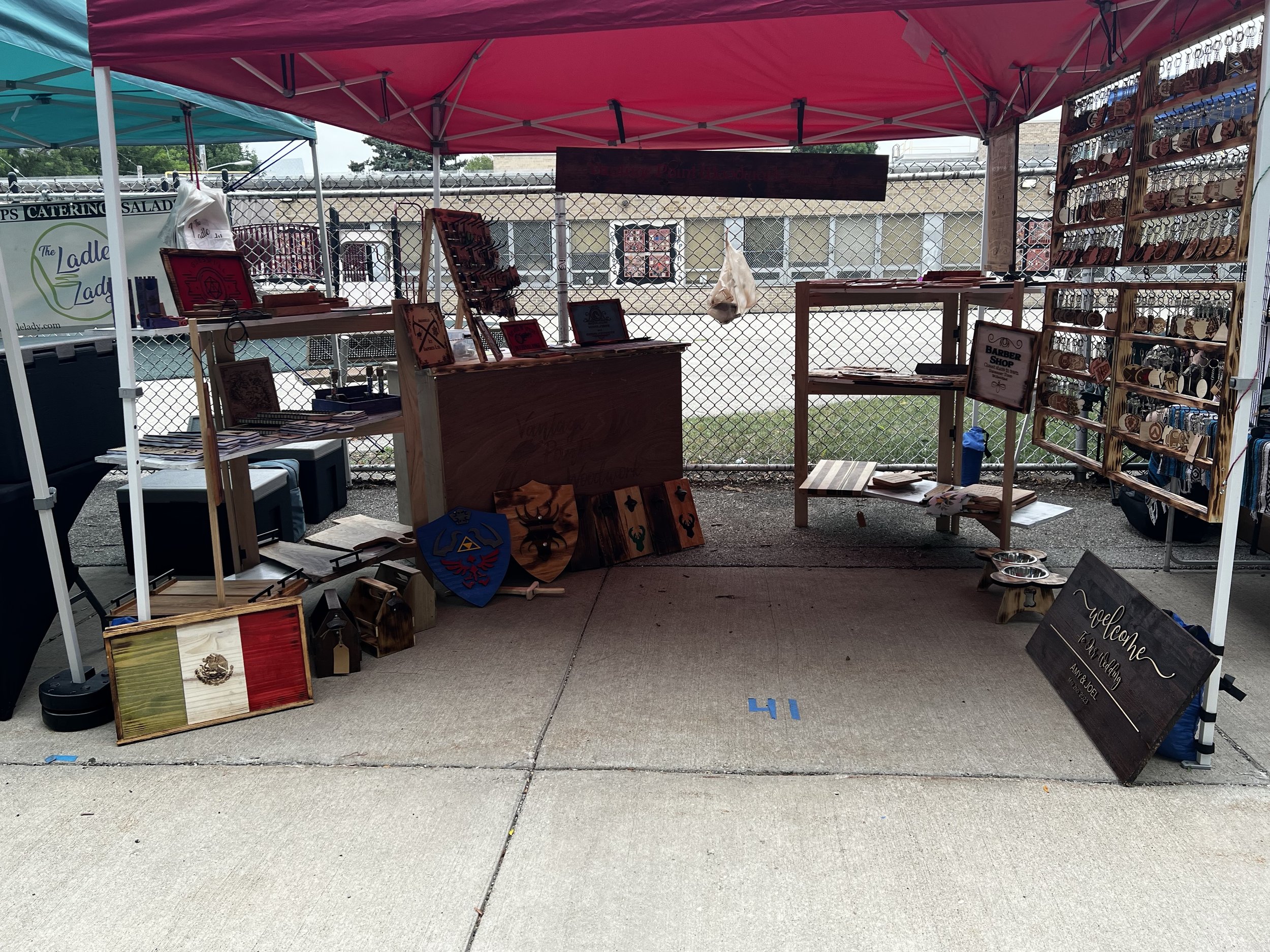 Outdoor market stall under a canopy displaying handcrafted wood items. Items include wooden plaques, shields, and keychains. The setting shows a concrete floor and chain-link fence in the background.