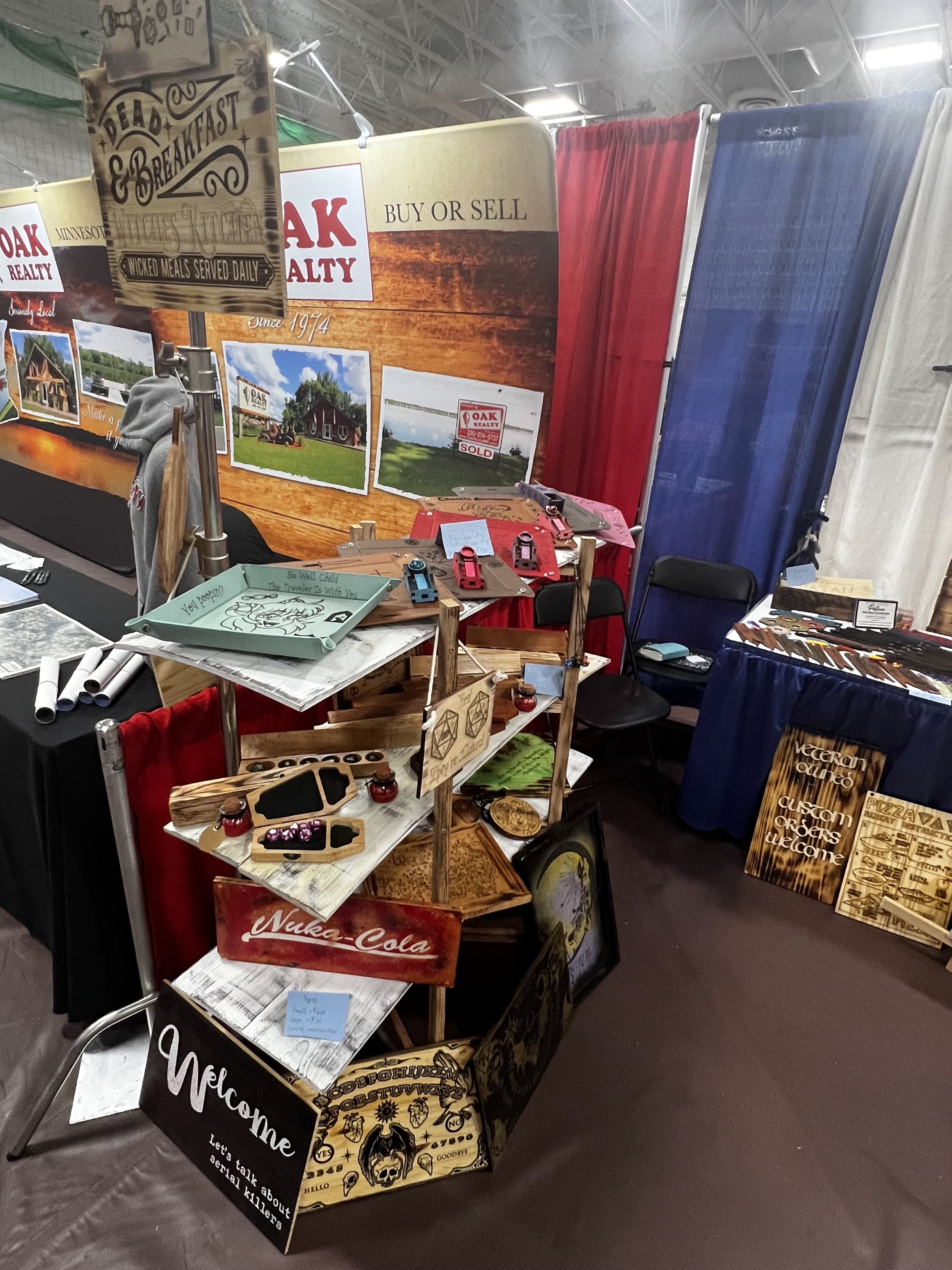 Trade show booth with wooden signs and plaques, featuring Oak Realty promotional materials and custom engraved art, including Nuka-Cola and Welcome designs, displayed on a multi-tier rack with colorful backgrounds.