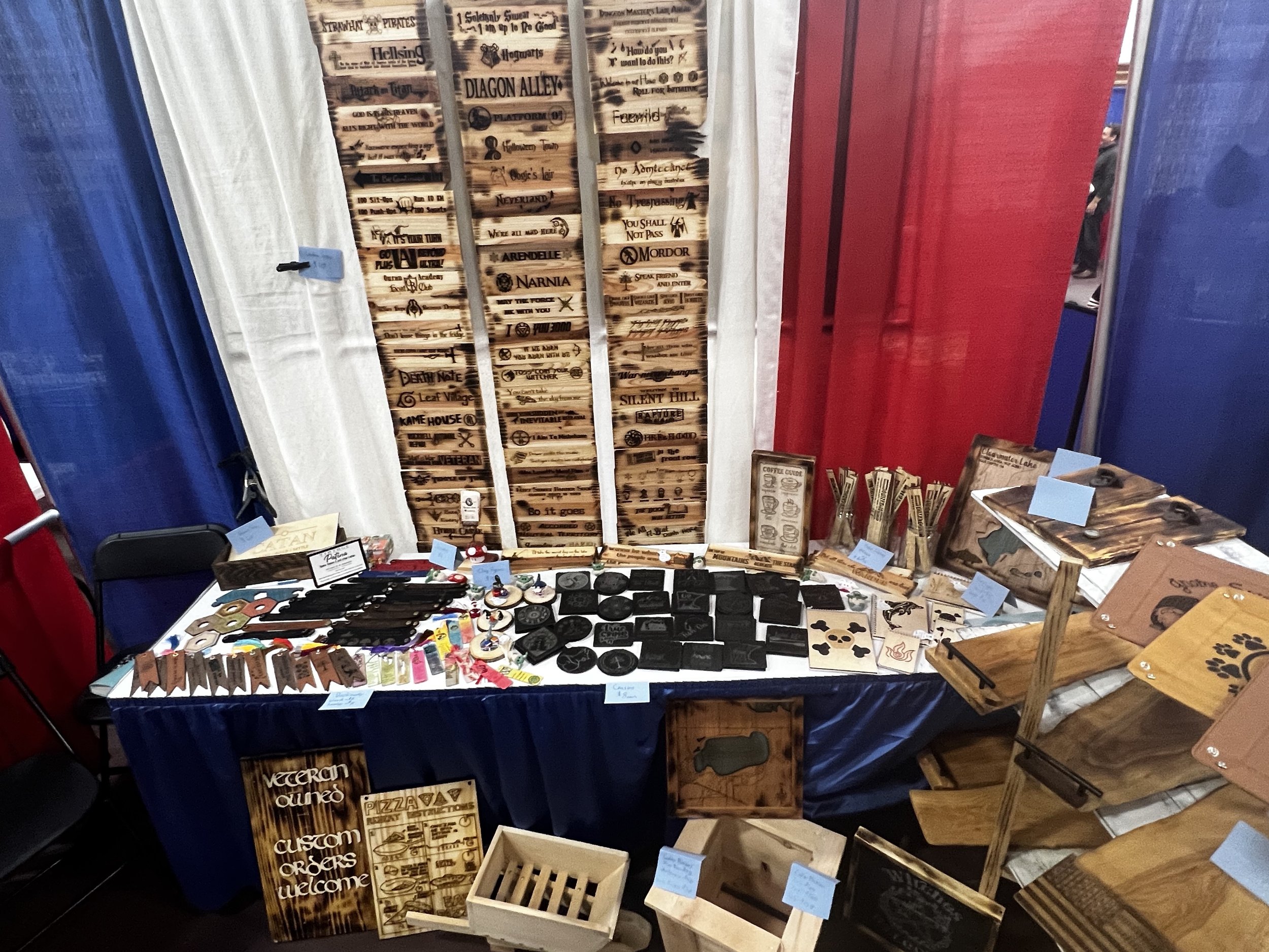 Booth at a craft fair displaying various wooden crafts and signs with engraved text and designs, including directional signs, coasters, and cutting boards. The booth is decorated with red, white, and blue curtains.