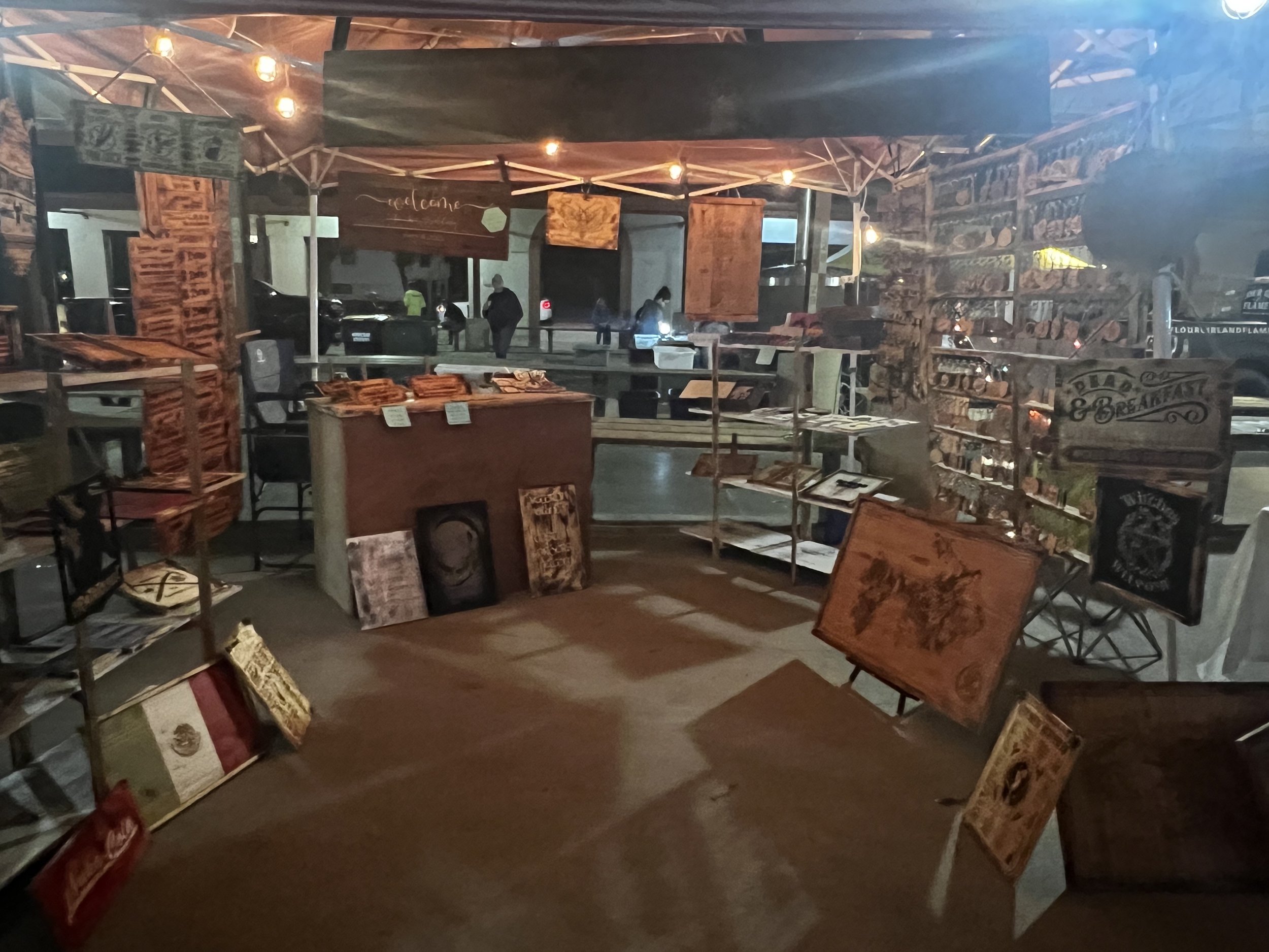 Outdoor art booth with framed artwork and decorative signs displayed, illuminated by string lights.