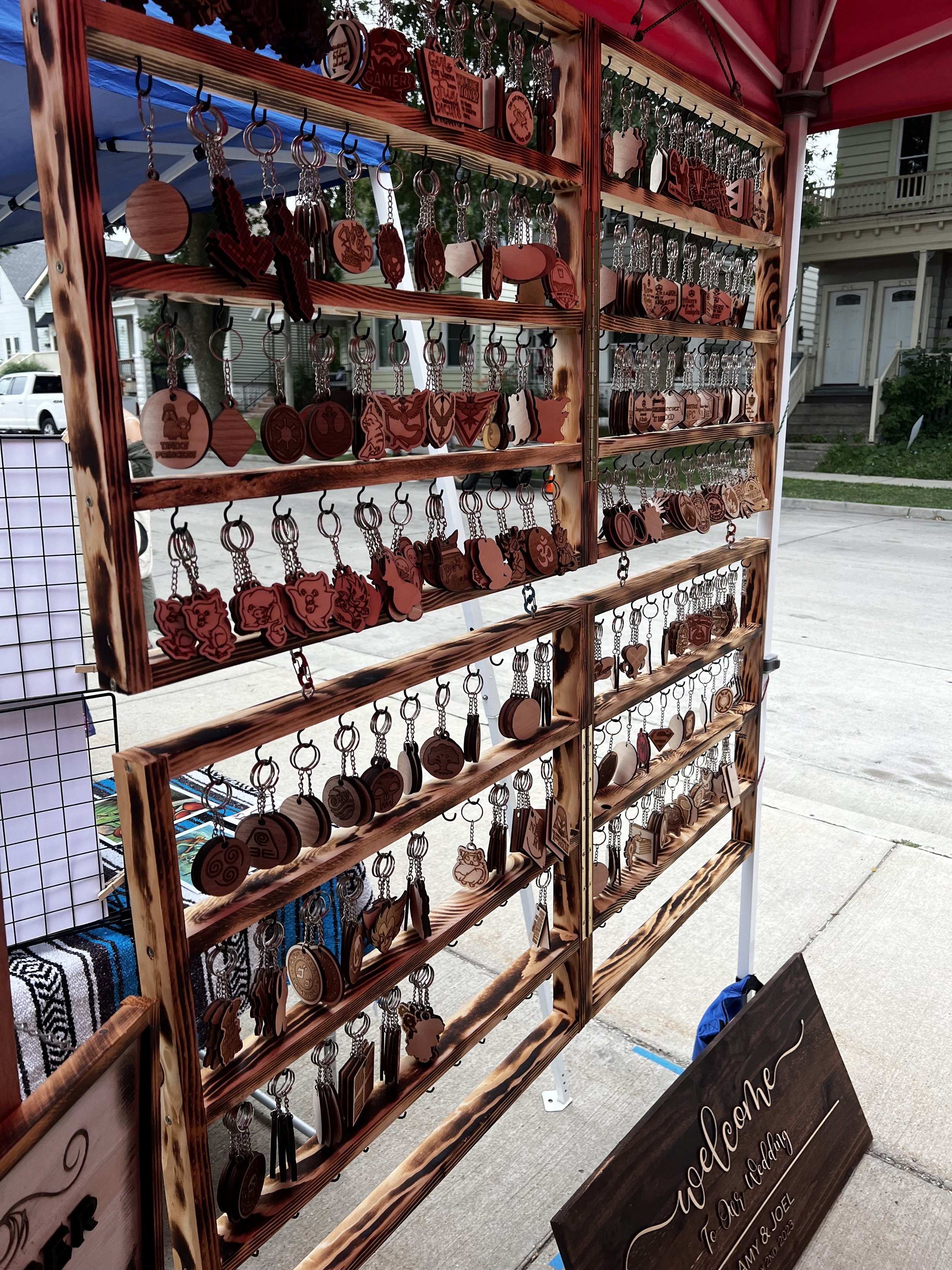 Wooden keychains displayed on a market stall with multiple rows of hooks under a red canopy. A wooden sign at the bottom reads "Welcome to Our Wedding."