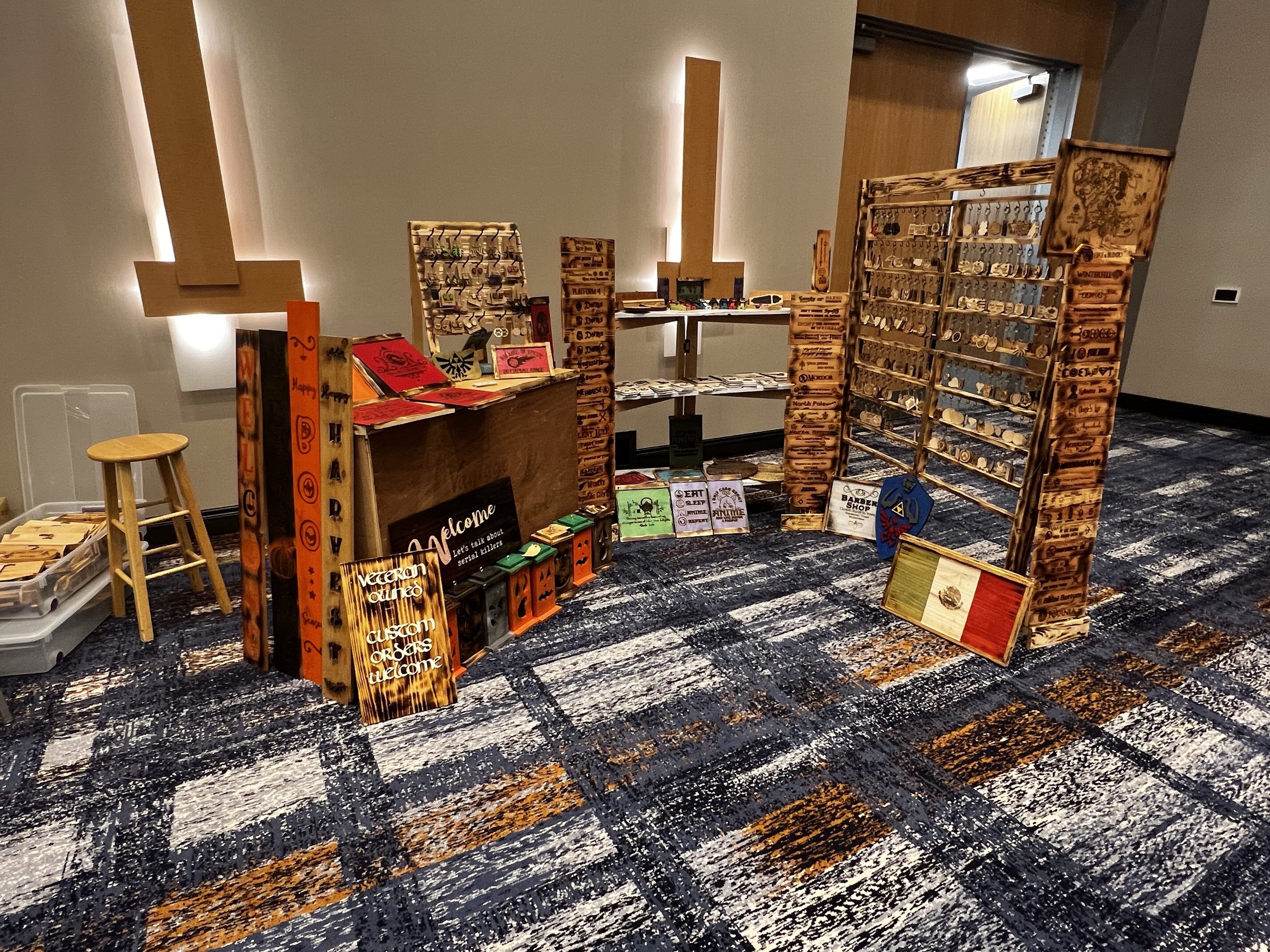 Craft fair booth displaying wooden signs and keychains on racks, with additional items on a table. Some signs feature holiday and welcome messages. A stool and storage bin are nearby.