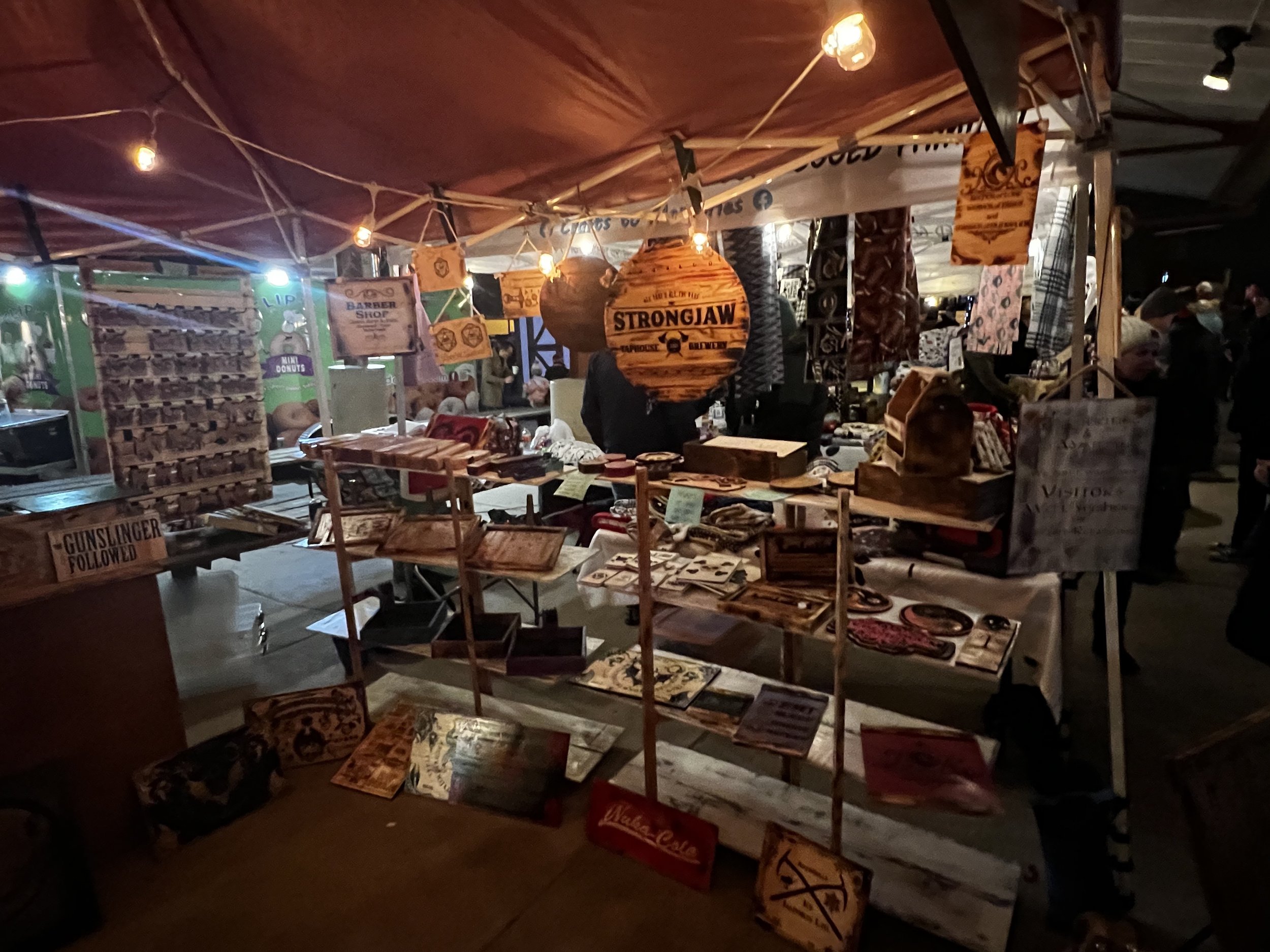 Outdoor market stall selling handmade crafts, wooden signs, and decorative items under a canopy tent, with various hanging lights and people browsing.