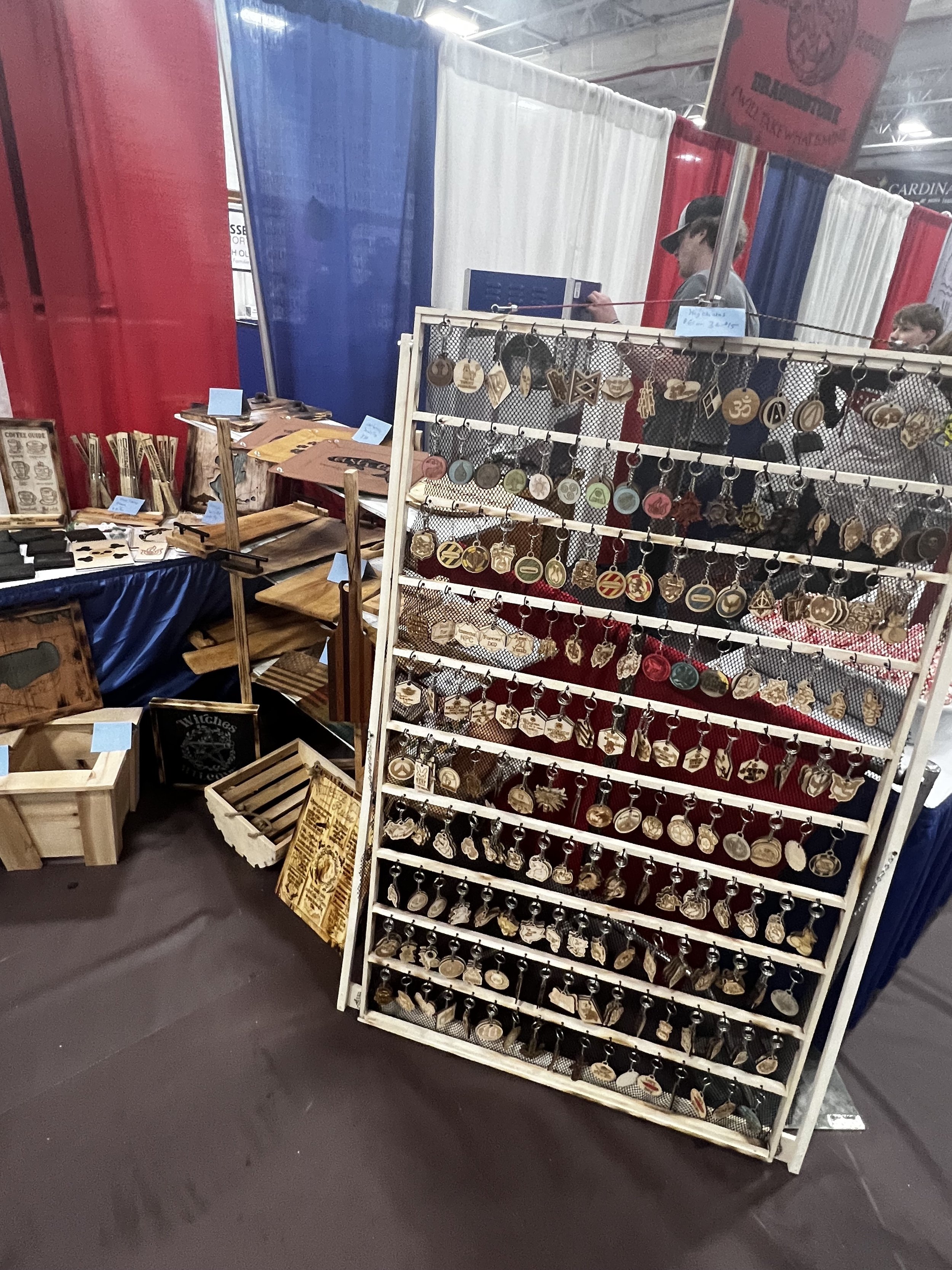 Display of handcrafted keychains with various designs, set up in a market stall with red, blue, and white curtains, featuring wooden items on tables and crates.