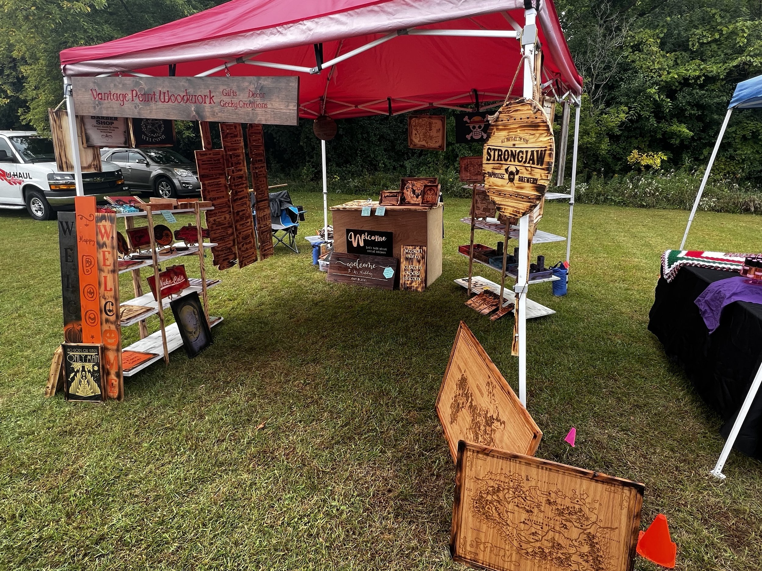 Outdoor craft fair booth under a red canopy, displaying various wooden crafts including signs, decor items, and maps. Visible text includes 'Vantage Point Woodwork' and 'Strongjaw.' The booth is set up on grass with surrounding vehicles and trees.