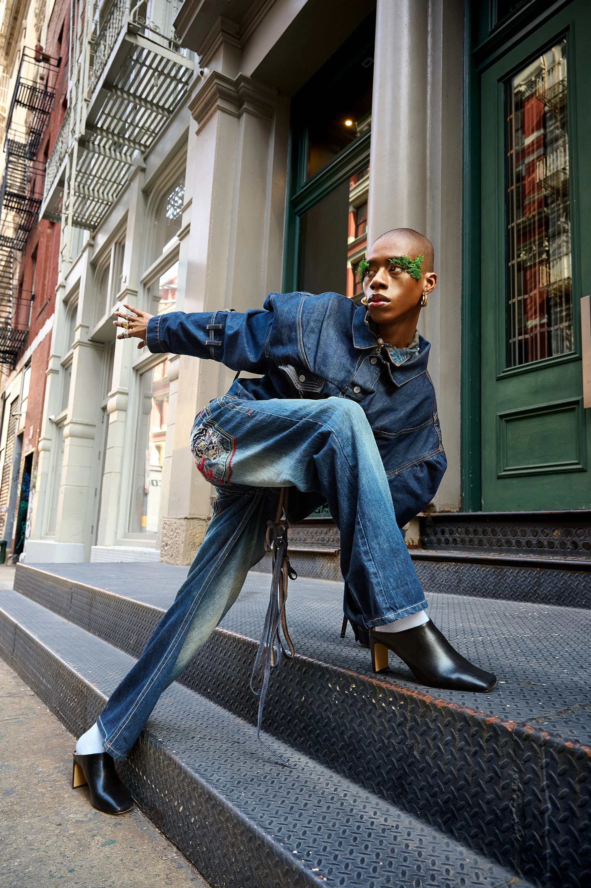A woman with a shaved head and green foliage around her eyes squatting on a staircase, wearing a denim jacket, wide-leg jeans, and black high-heeled boots, in front of a city building.