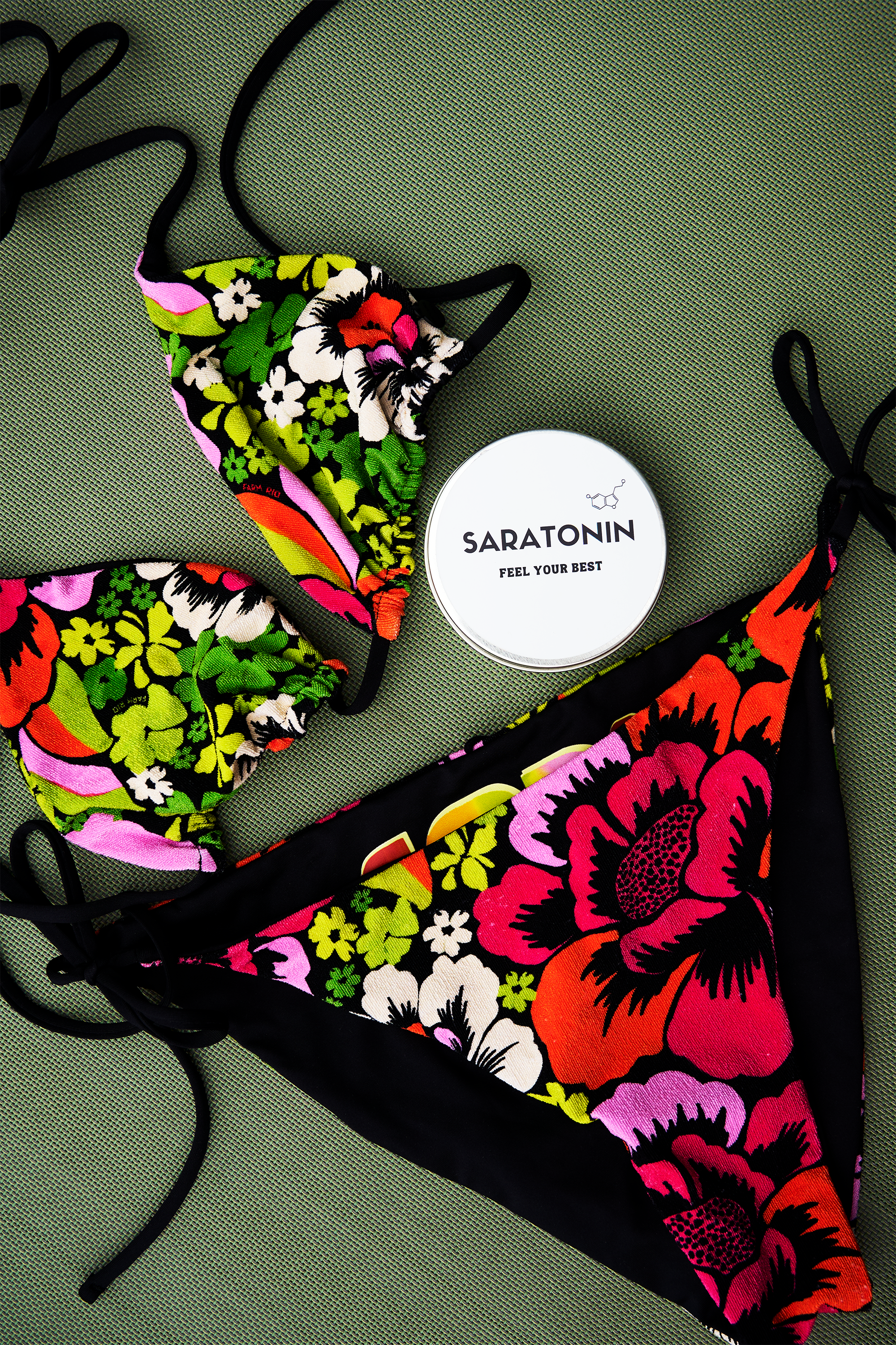 Colorful floral patterned face mask with black straps and a round white container labeled 'SARATONIN FEEL YOUR BEST' on a green textured surface.
