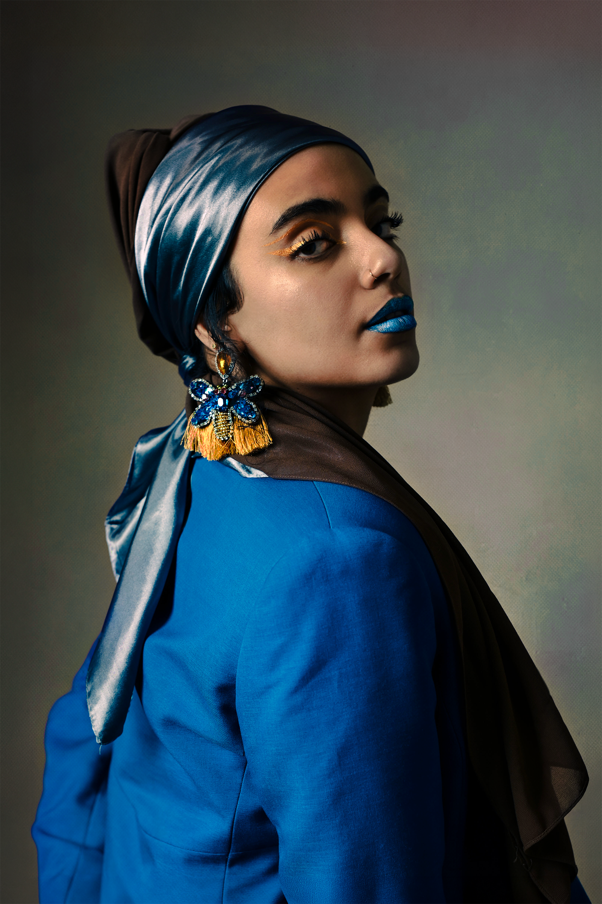 A woman with a shiny blue headscarf, large gold and blue beaded earrings with a bee design, and blue lipstick. She is dressed in a blue blazer and has makeup with gold eyeliner, against a dark background.