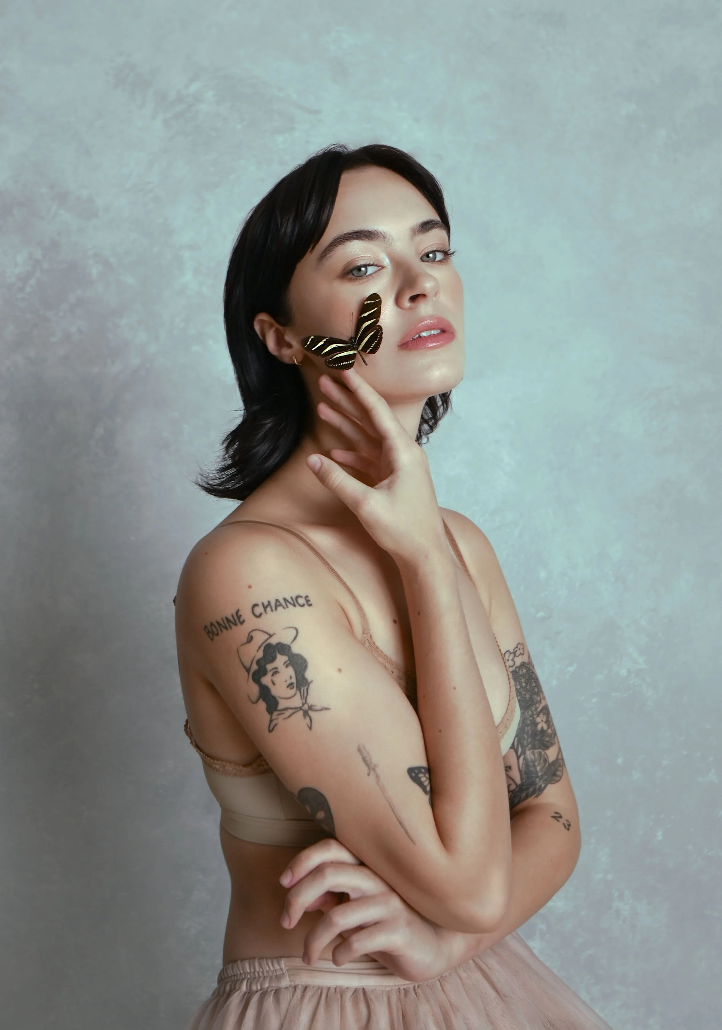 A woman with black hair and tattoos on her arm is holding her neck with her right hand. She has a butterfly on her left cheek and is wearing minimal clothing. The background is a pastel-colored, textured surface.