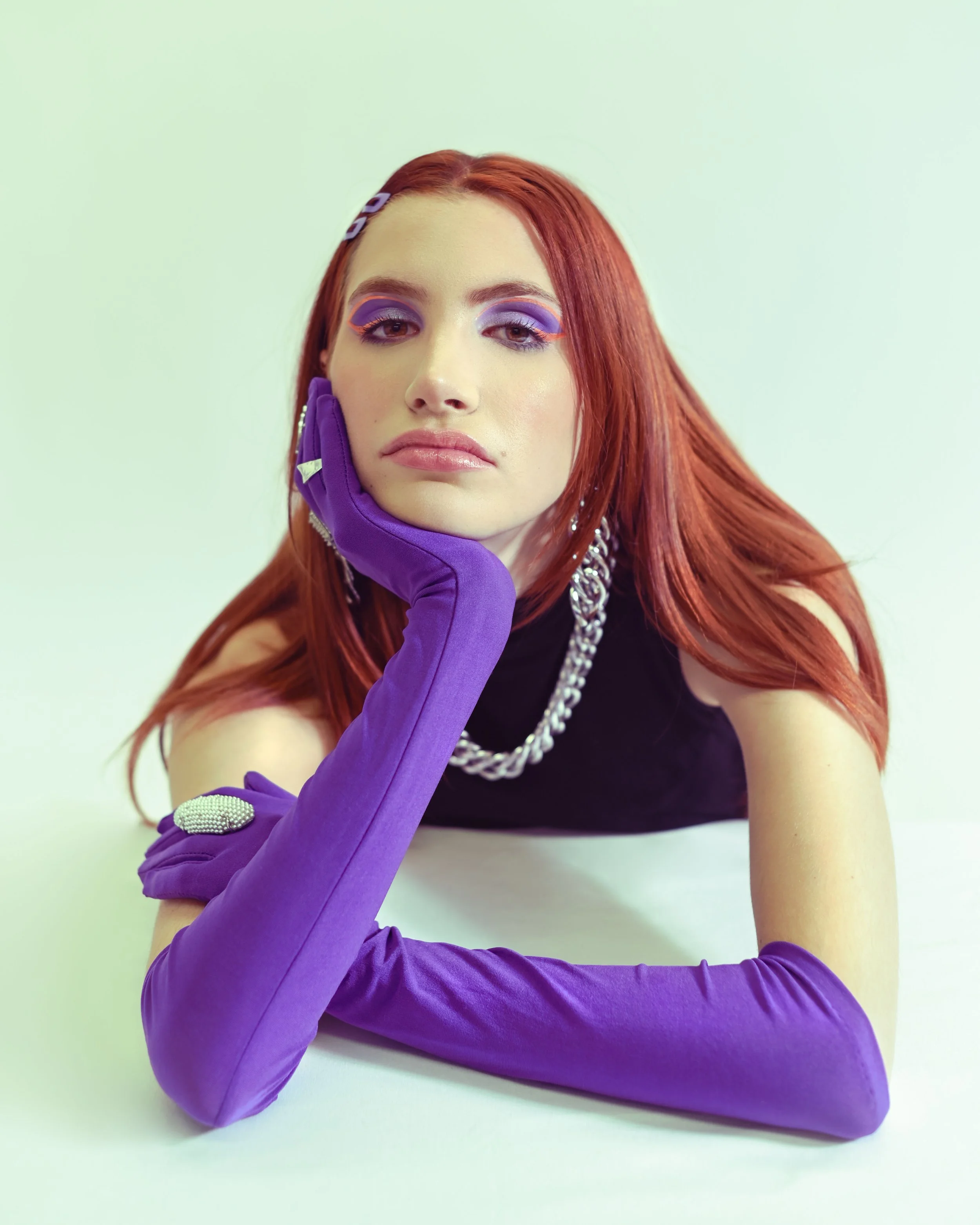 A woman with vibrant red hair and purple gloves lying on a white background, styled with makeup, jewelry, and hair accessories.