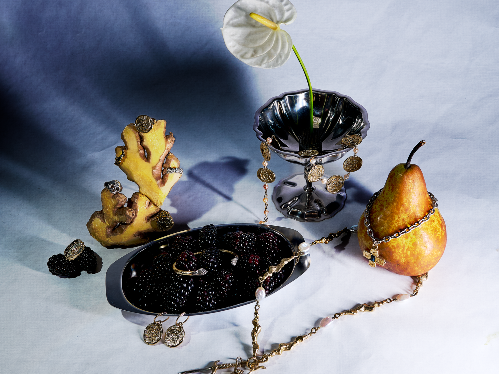A still life arrangement of jewelry pieces displayed alongside fruits, including a pear, pear slice, and a pomegranate, with a decorative vase holding an anthurium flower.