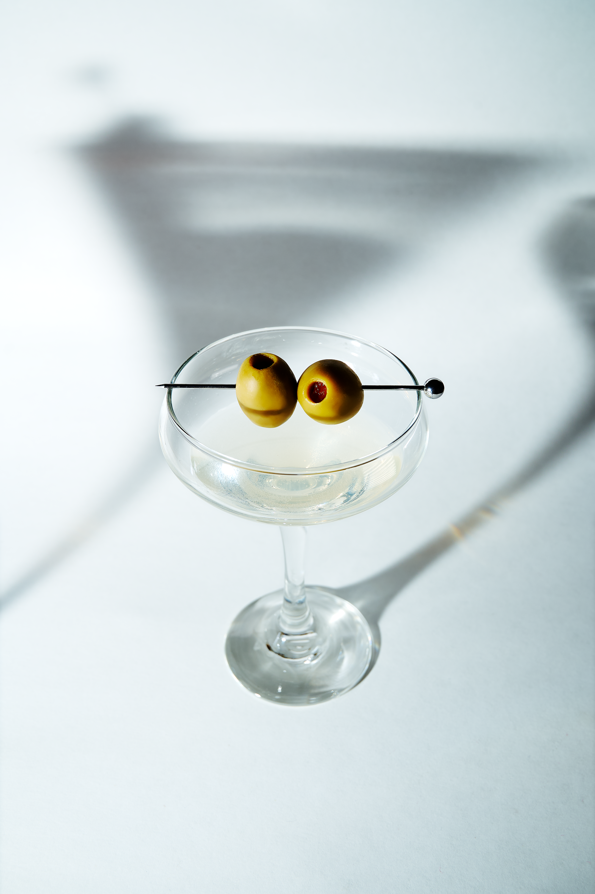 A martini glass with two green olives on a skewer, set against a white background, with shadows cast on the surface.