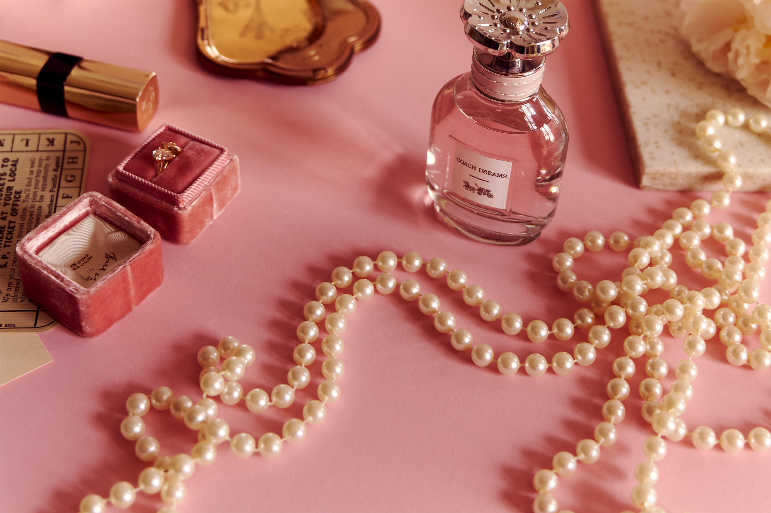 A pink surface with jewelry and perfume items, including pearl necklaces, a ring in a pink velvet box, and a bottle labeled 'Coach Dreams' surrounded by a pearl necklace, alongside a slice of cake with frosting and pearls, and a ticket stub.