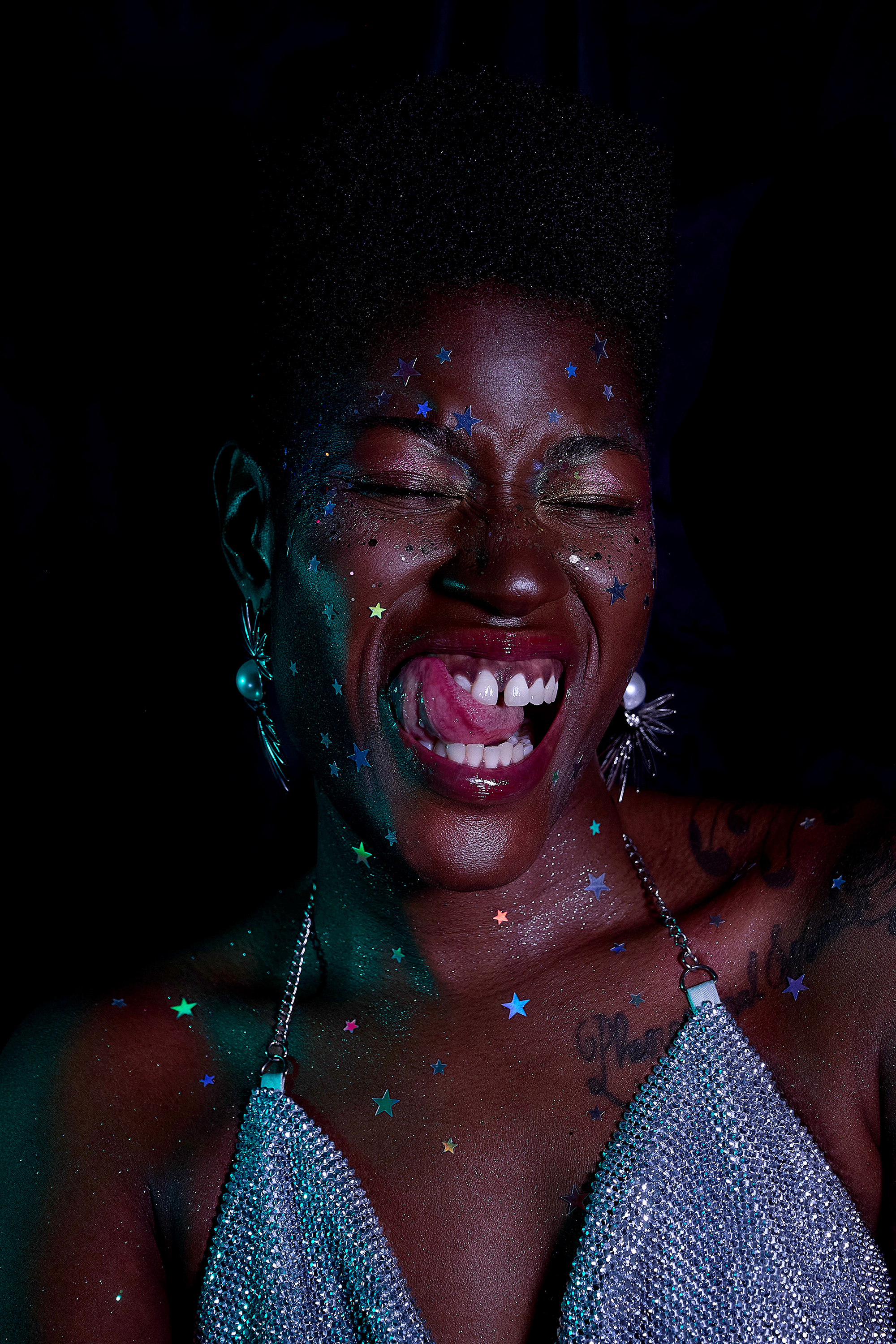 A woman with dark skin and short, curly hair sticking out of a black hat, smiling widely with her tongue sticking out, covered in colorful star-shaped confetti and glitter, wearing large earrings, a long necklace, and a sparkly dress.