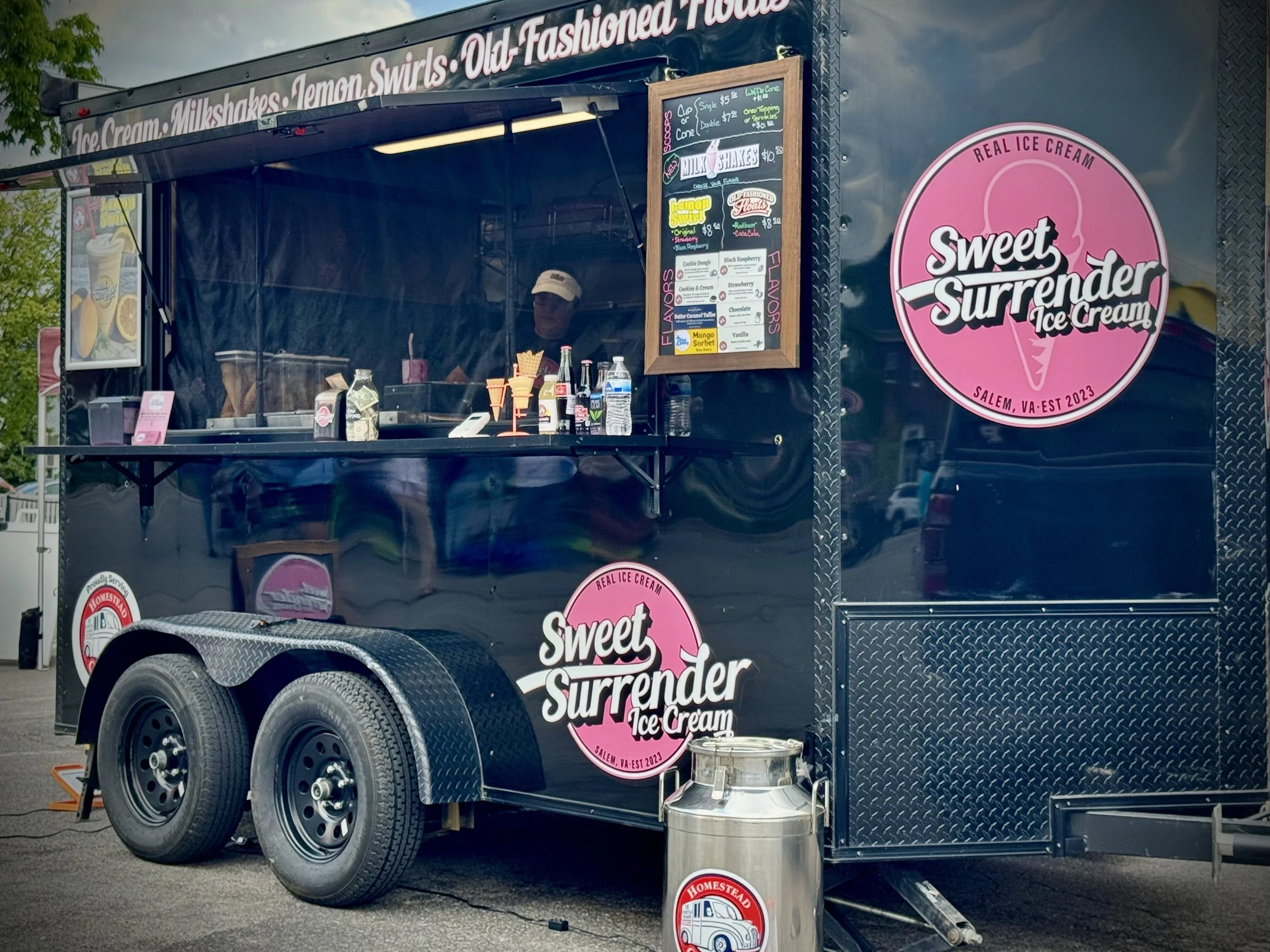 A black ice cream truck with pink and white branding for Sweet Surrender Ice Cream. The truck lists flavors like Lemon Swirl, Ruebber, Coca Cola, cookies and cream, strawberry, and more on a chalkboard menu, and has a large pink circular logo on the side. Several cones and bottled drinks are on the counter, with a person wearing a hat working inside. The truck is parked outdoors near trees and other structures.