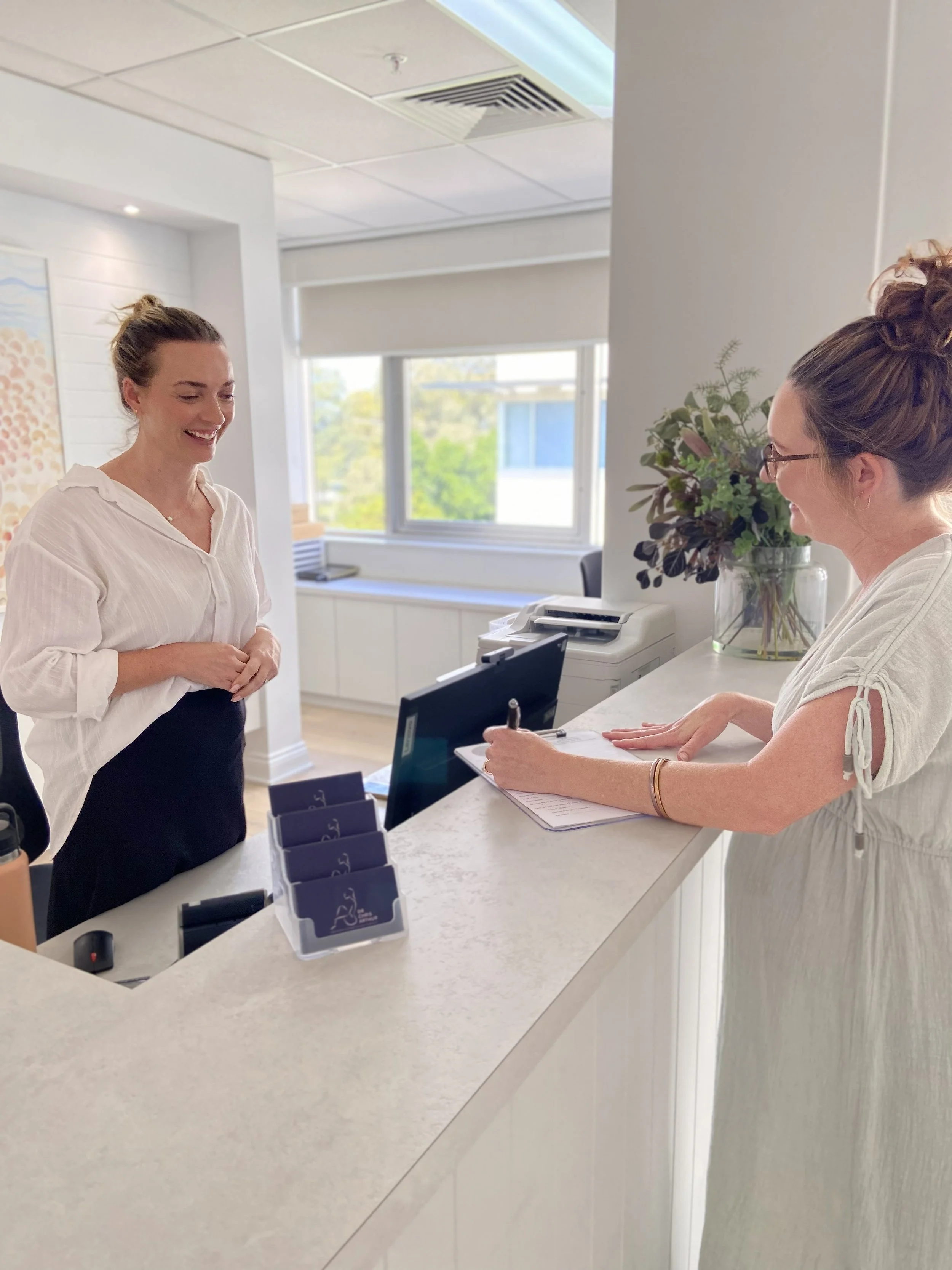 Two women working at a reception desk, with one standing and smiling, and the other sitting and writing on a notepad, in a modern office environment.