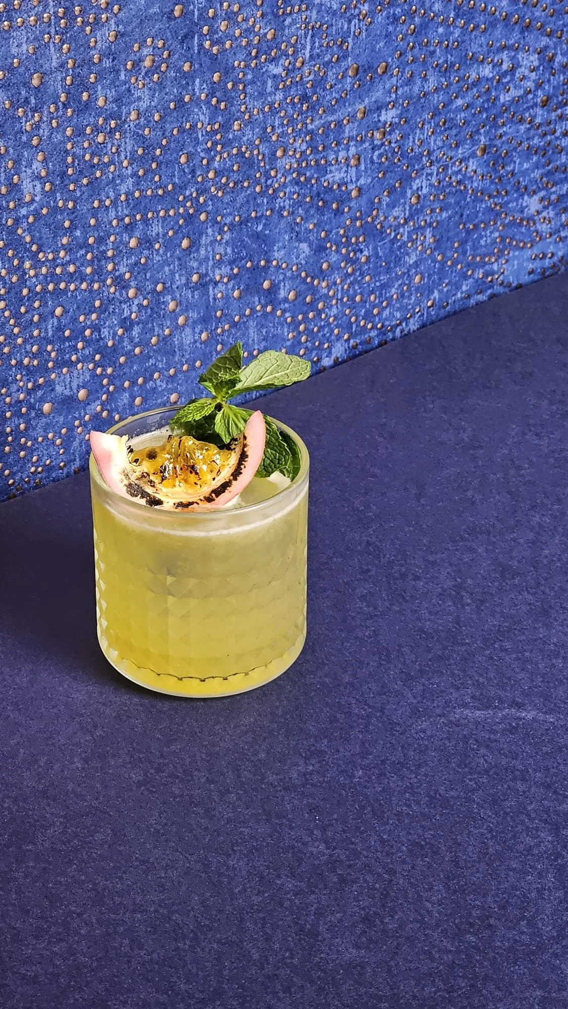 A yellow cocktail garnished with mint, a slice of pink dragon fruit, and passion fruit seeds, served in a textured glass on a blue surface with a decorative blue wall in the background.