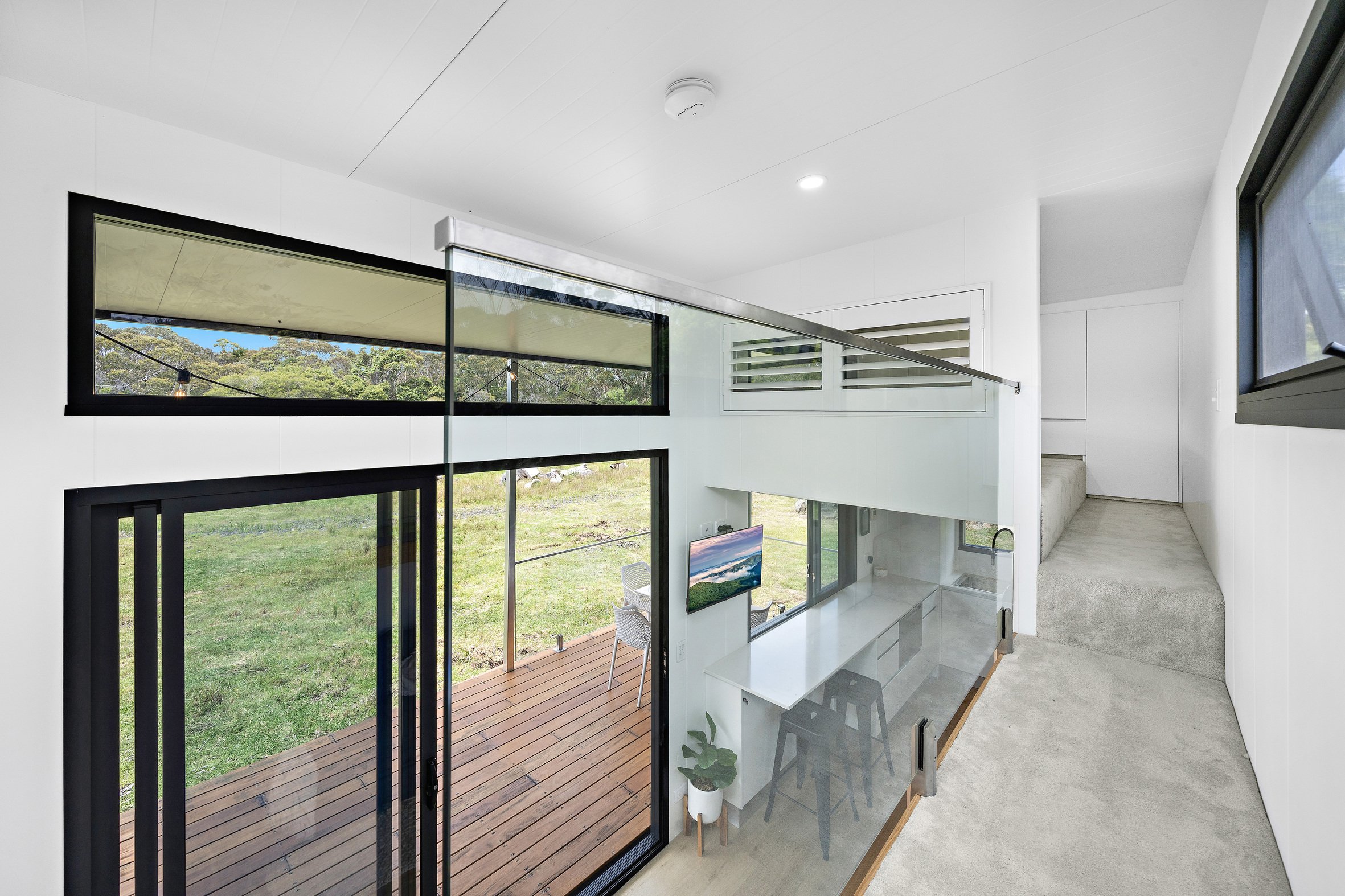 Modern interior with white walls and large windows overlooking a deck and grassy area. Contains a loft space with glass railing, a minimalistic workspace below, and a small plant. Natural light brightens the area.