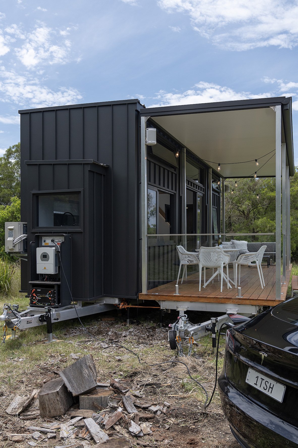 Black tiny house on wheels with a porch, featuring a trailer hitch and outdoor seating area, located in a natural setting with logs and greenery.