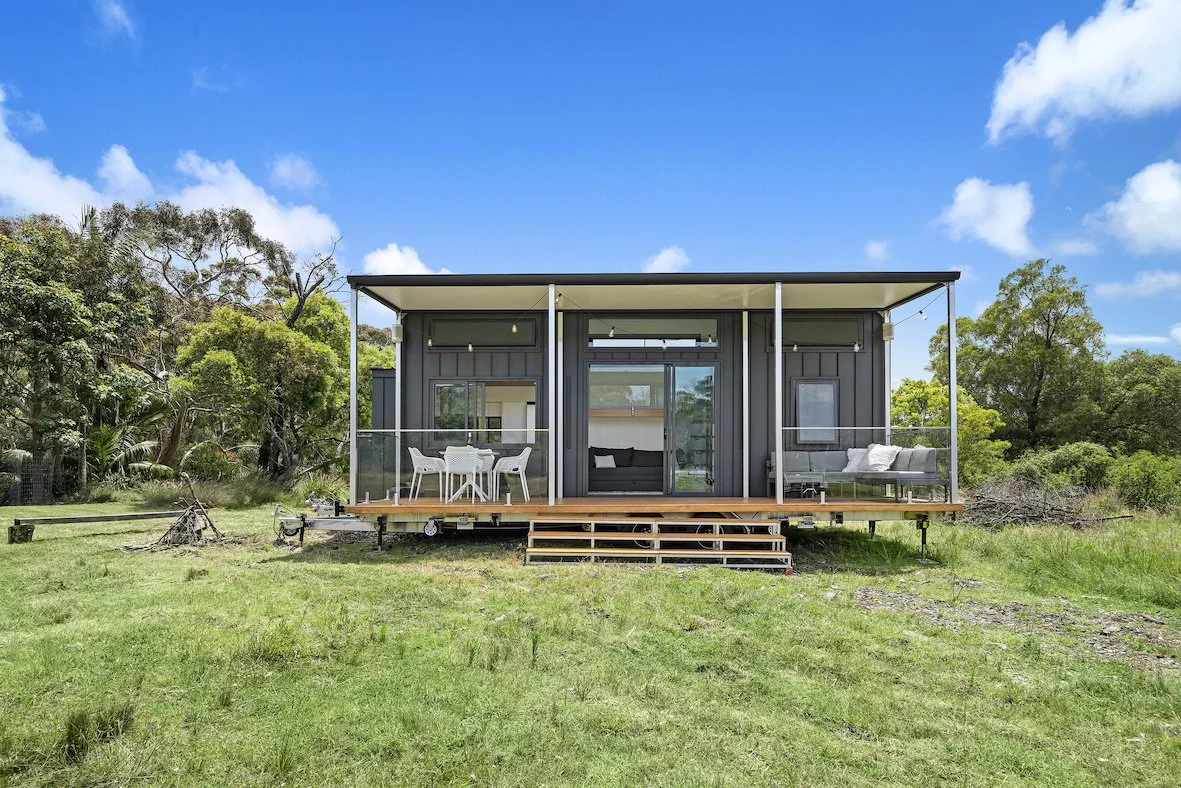 Small modern modular home with a wooden deck and outdoor seating, surrounded by greenery and trees, under a blue sky.