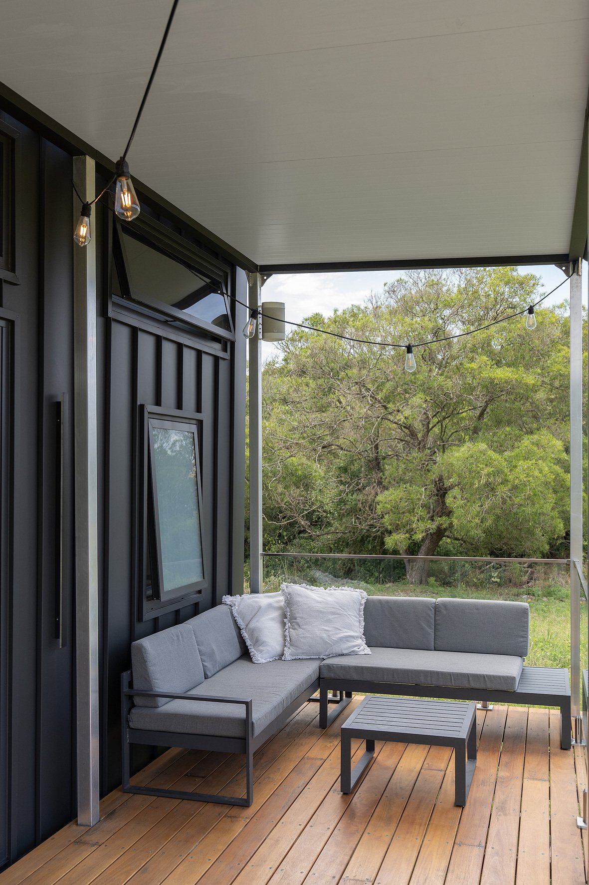 Modern outdoor patio with gray sectional sofa, wooden decking, string lights, and view of trees.