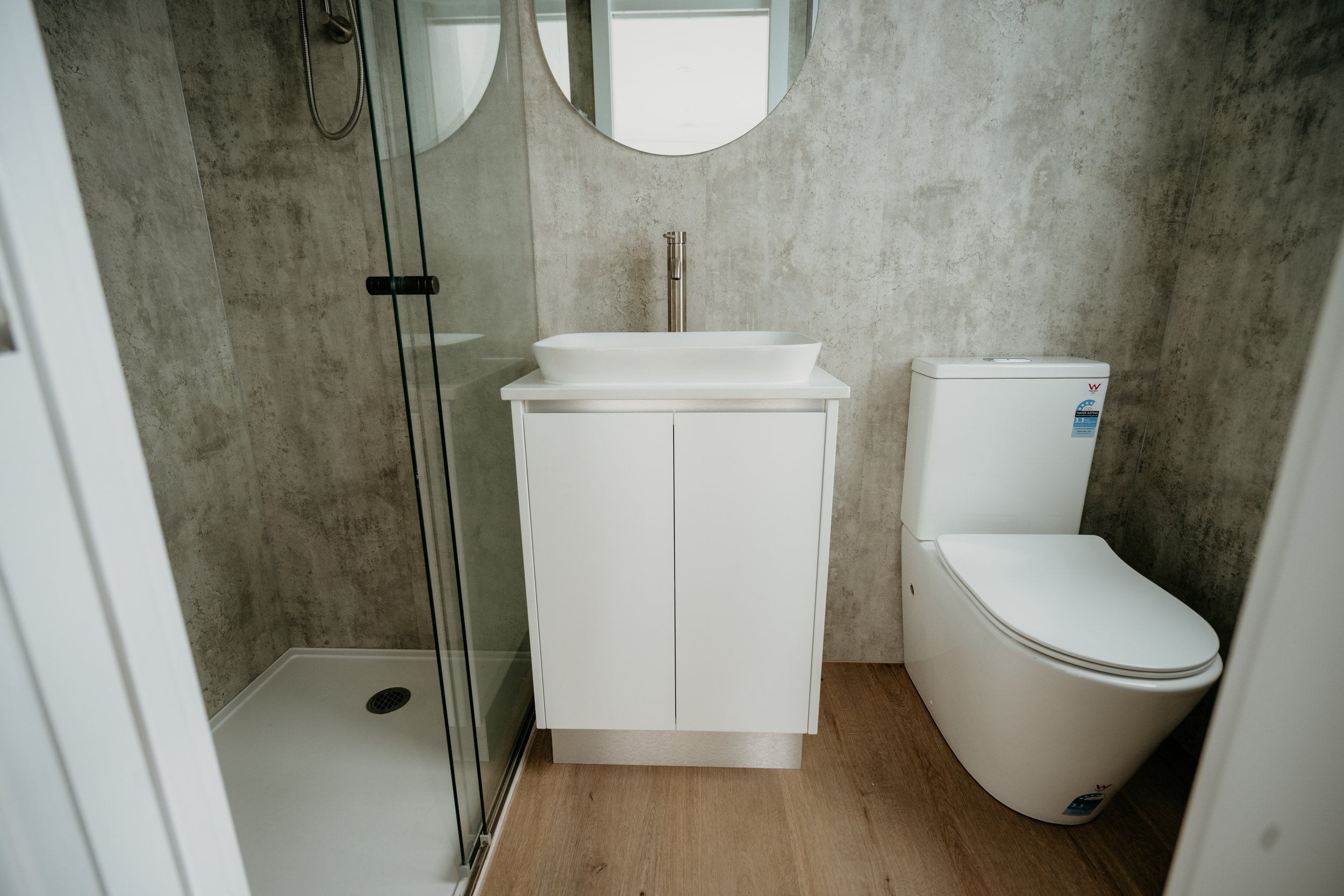 Modern bathroom with glass shower, white sink on cabinet, round mirror, and toilet on wood floor.