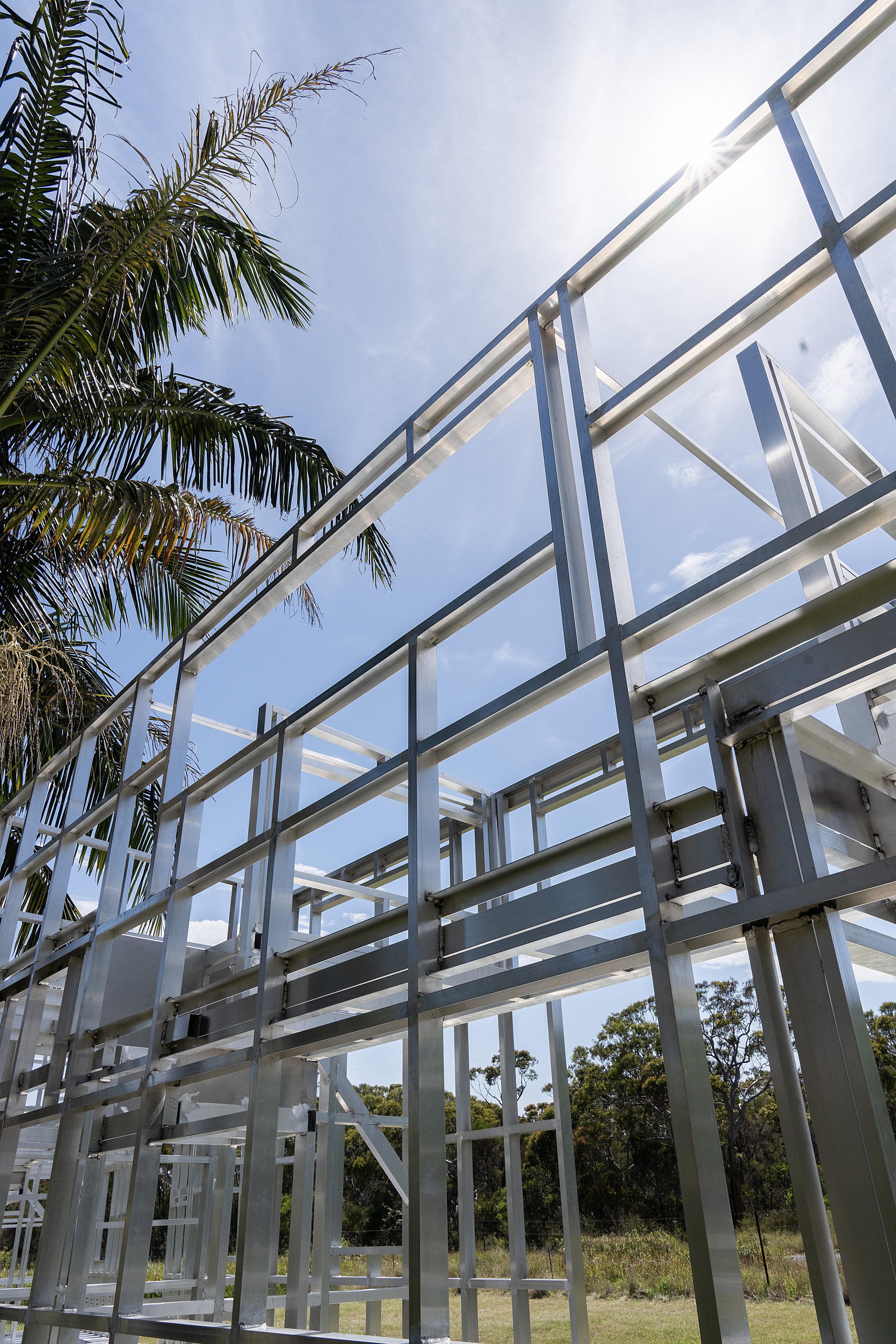Steel frame structure under construction with palm tree and bright sun in background.