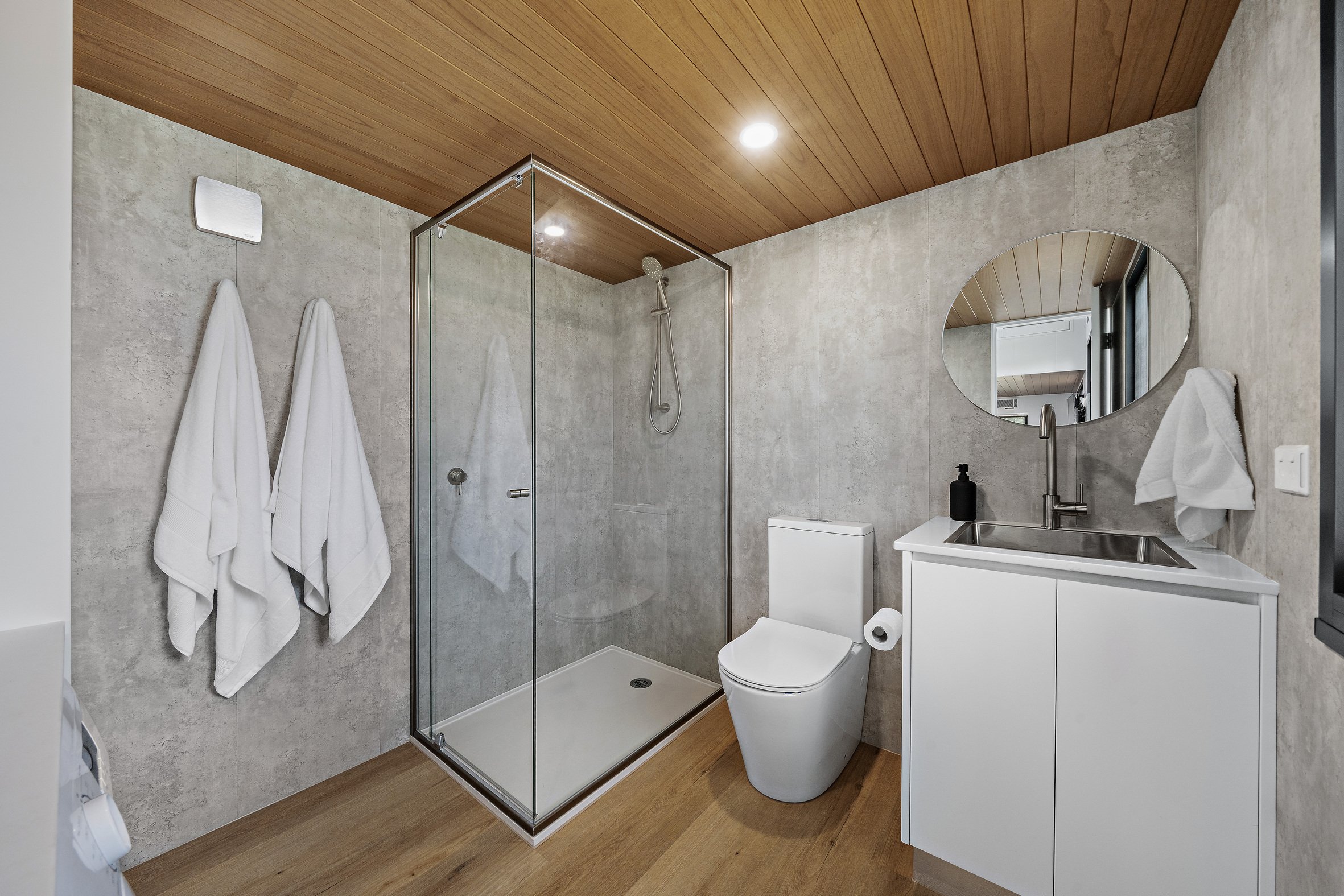 Modern bathroom with a glass shower, white toilet, vanity with a round mirror, and white walls. Two white towels hang on the wall, and a wooden ceiling contrasts with the minimalist design.