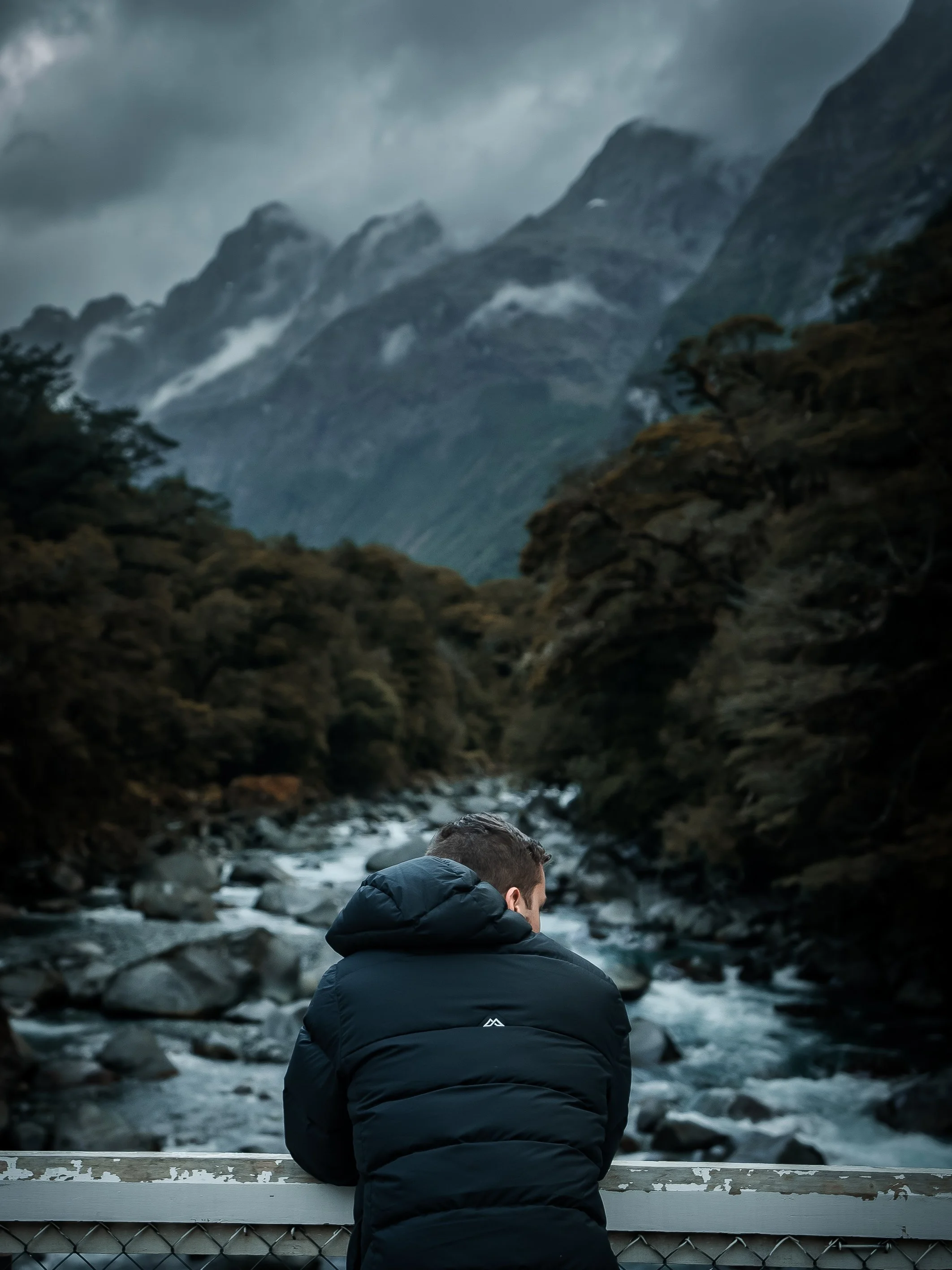 A person wearing a black puffer jacket is sitting on a bridge railing, looking out over a river flowing through a mountainous landscape with cloudy skies.