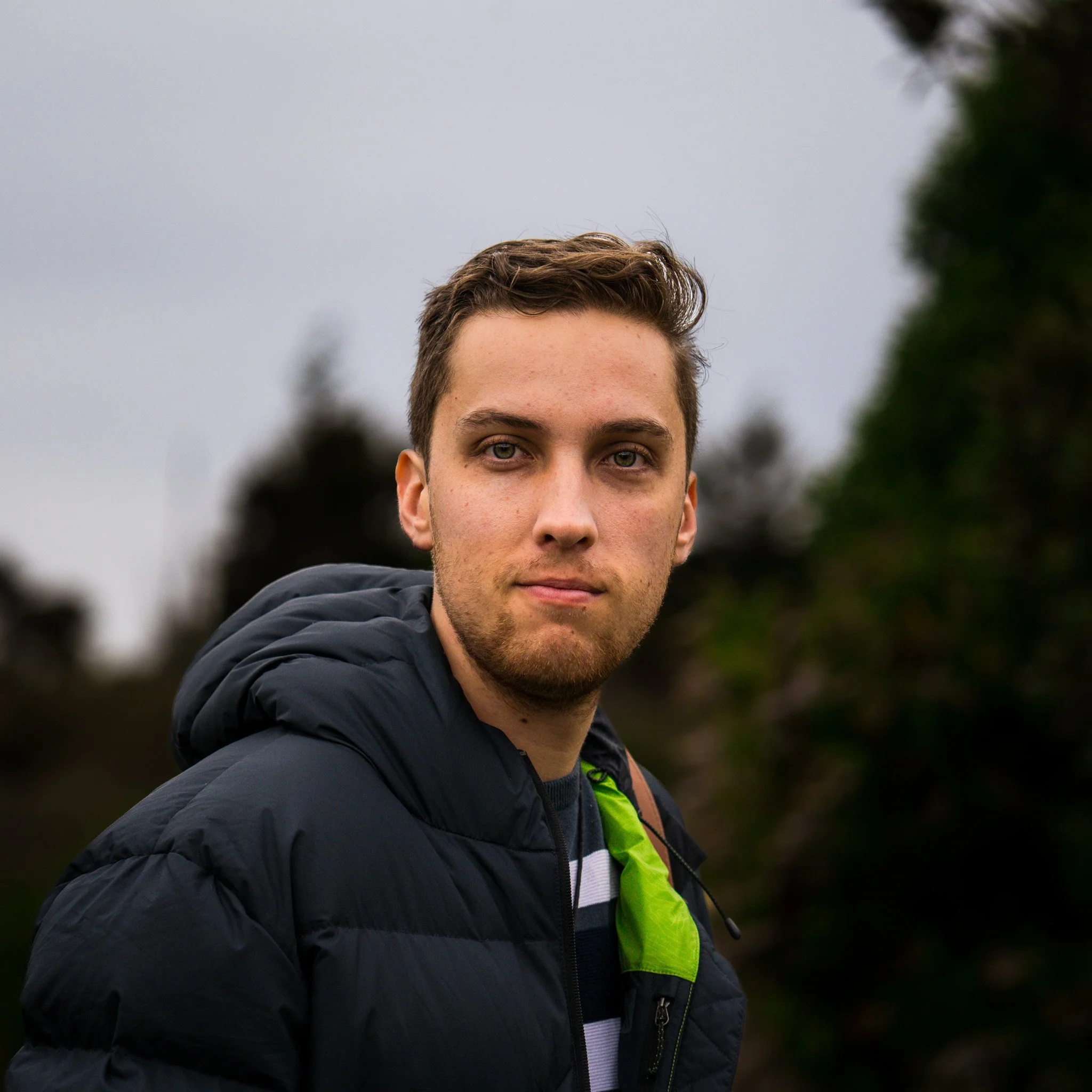 A young man with short brown hair and a light beard, wearing a dark puffer jacket and a striped shirt, standing outdoors on a cloudy day with blurred trees in the background.