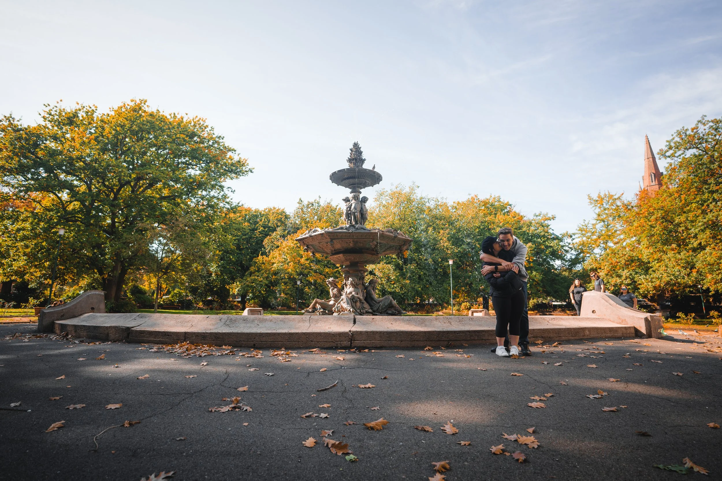 People hugging near a fountain in a park during fall, with trees showing autumn colors and a church steeple in the background.